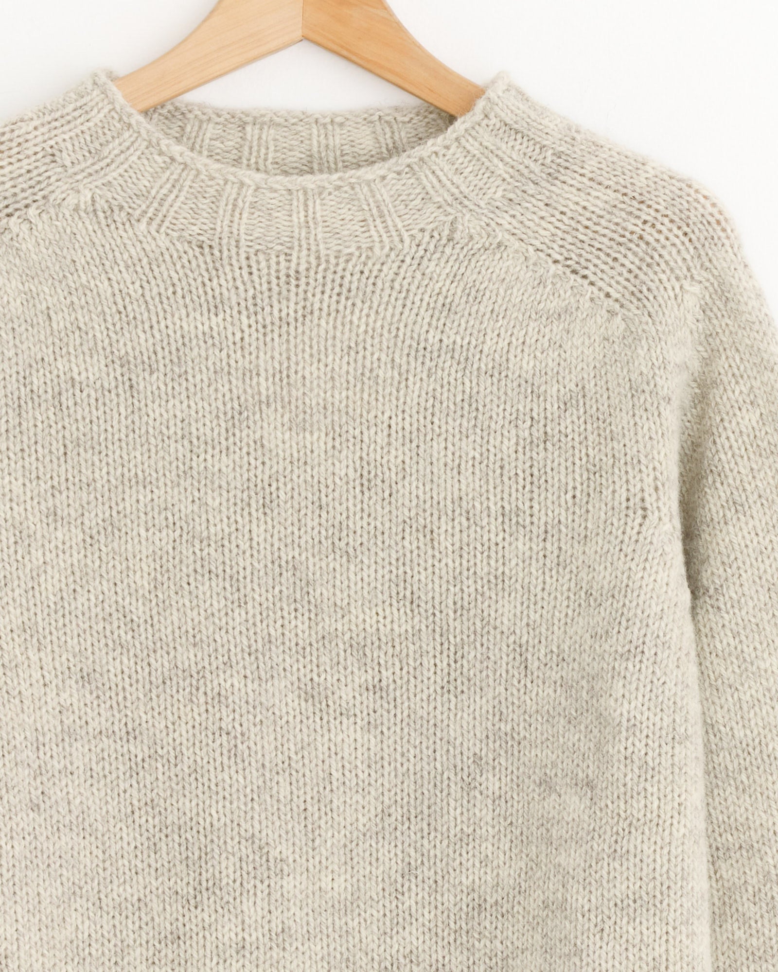 A close-up of the MHL by Margaret Howell Chunky Seamless Crewneck in Natural, featuring beige knit wool and a ribbed neckline, displayed on a wooden hanger. For fit and sizing help, our online chat is available.