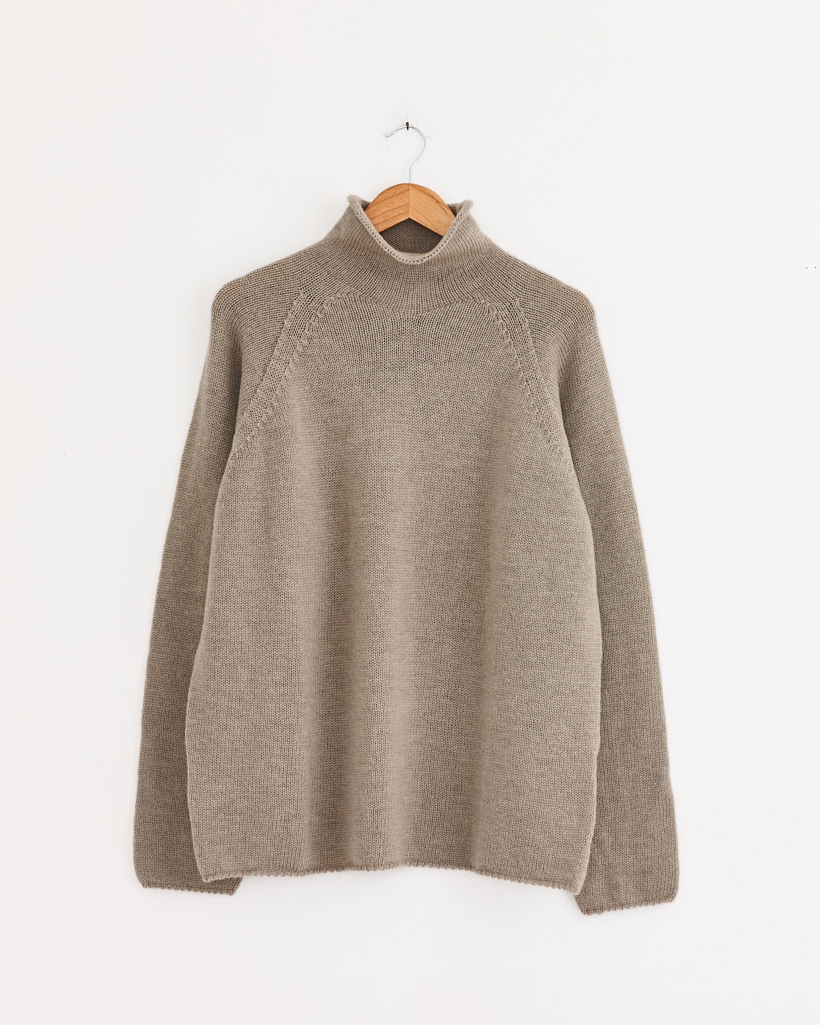 The Rolled Edge Roll Neck in Taupe by MHL by Margaret Howell, a long-sleeve knit sweater made from Japanese wool, features a mock turtleneck and hangs on a wooden hanger against a white wall.