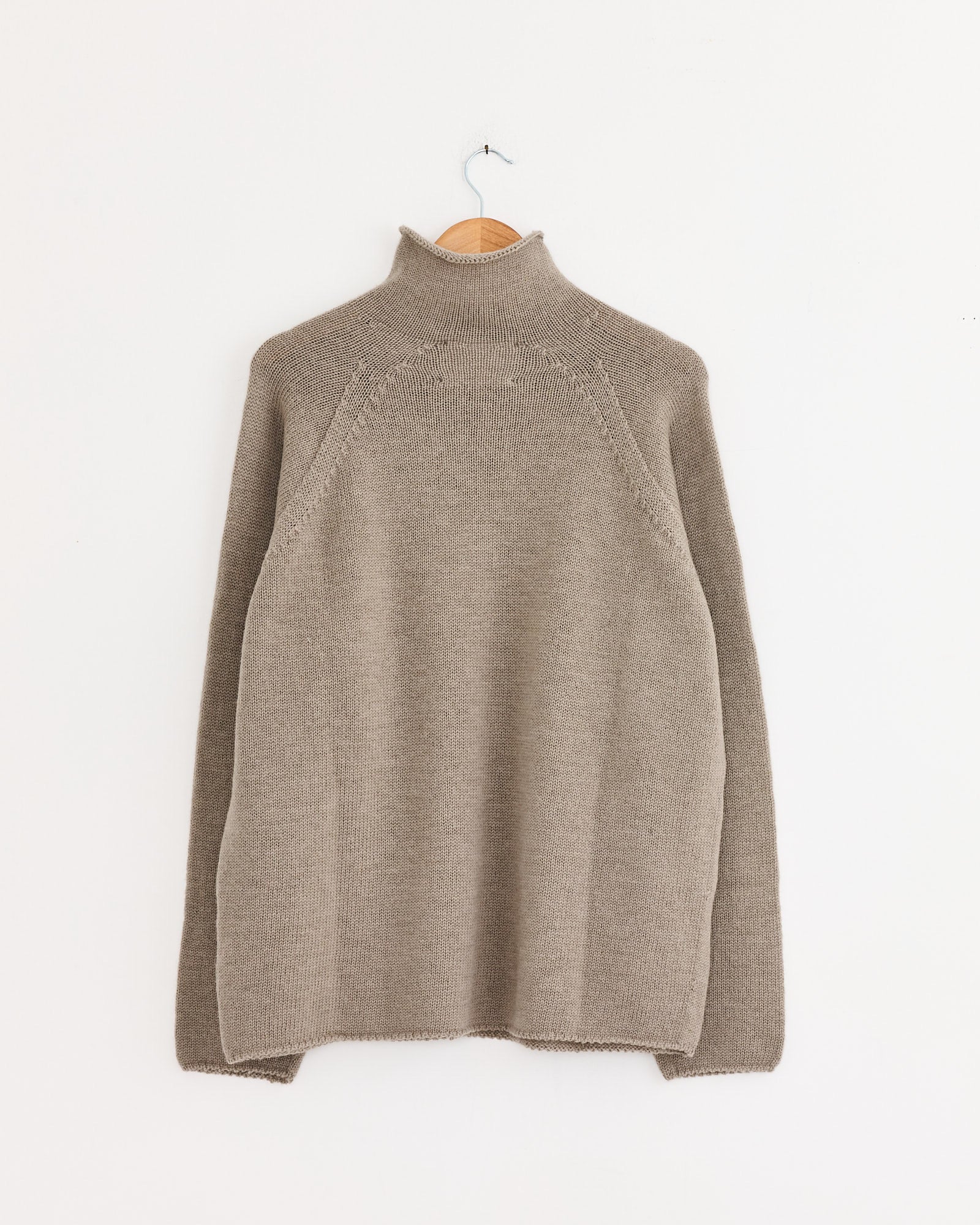 The Rolled Edge Roll Neck in Taupe by MHL by Margaret Howell, a long-sleeve Japanese spun wool sweater, hangs on a wooden hanger against a plain white wall.