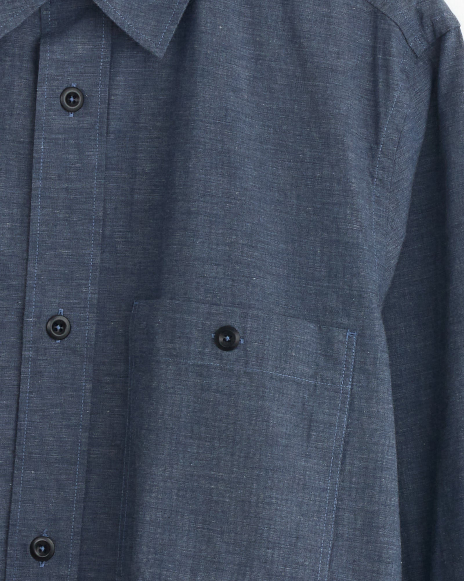 Close-up of the Overall Shirt in Uniform Blue by MHL by Margaret Howell, featuring the collar, front buttons, and a buttoned chest pocket. For fit or sizing questions, contact our online chat team.