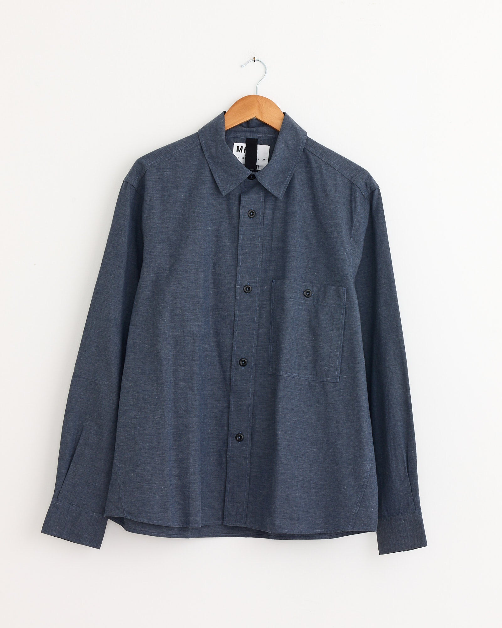 The Overall Shirt in Uniform Blue by MHL by Margaret Howell, a long-sleeve button-up with a chest pocket, hangs on a wooden hanger against a white wall. For sizing or fit questions, contact us via online chat.
