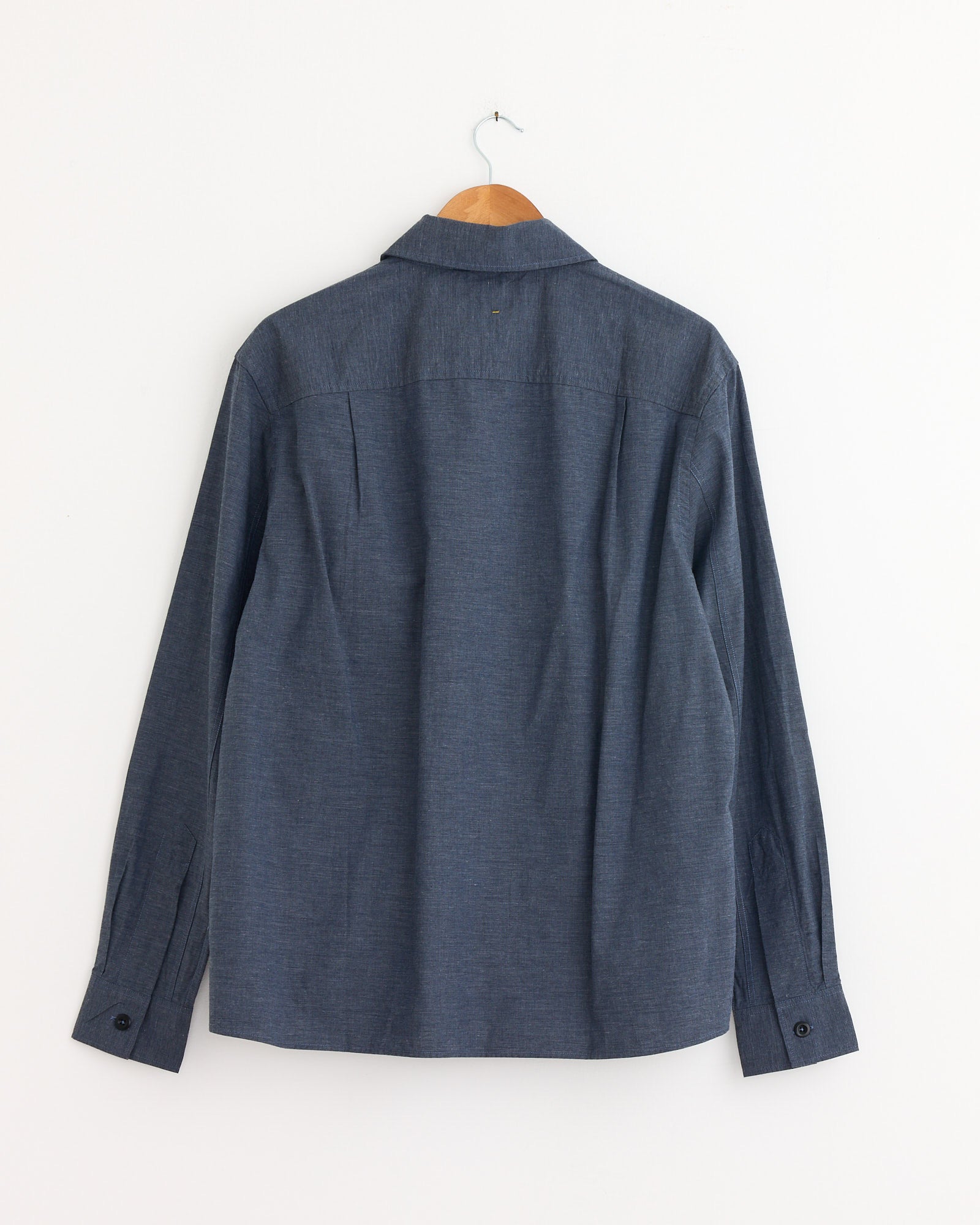 The MHL by Margaret Howell Overall Shirt in Uniform Blue, a long-sleeve button-up denim style, hangs on a wooden hanger with the back facing forward—ideal for checking sizing or fit through our online chat.