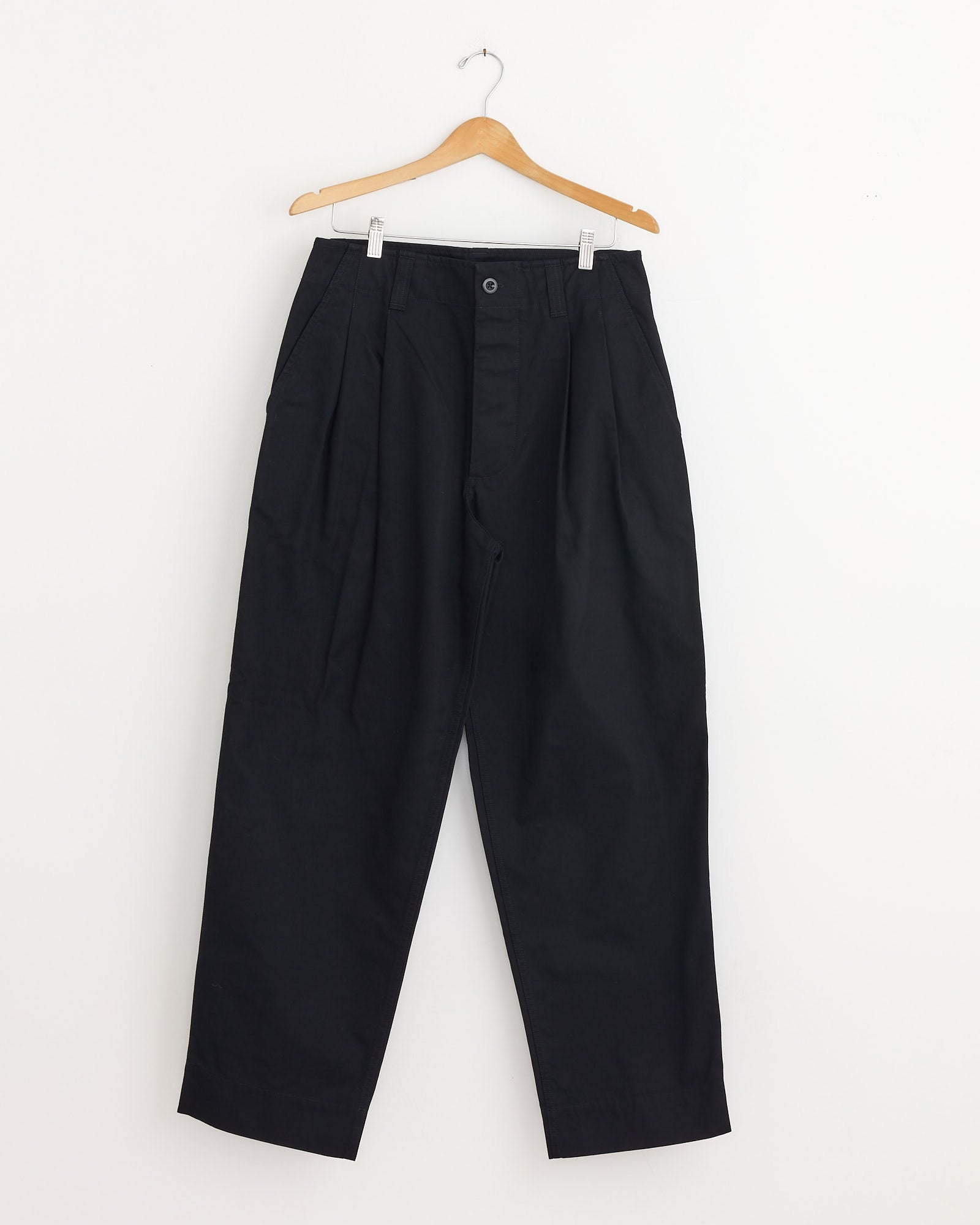 The Stitch Pleat Trouser in Black by MHL by Margaret Howell, featuring loose pleats, hangs on a wooden hanger against a white wall. For sizing or fit assistance, chat with us online.