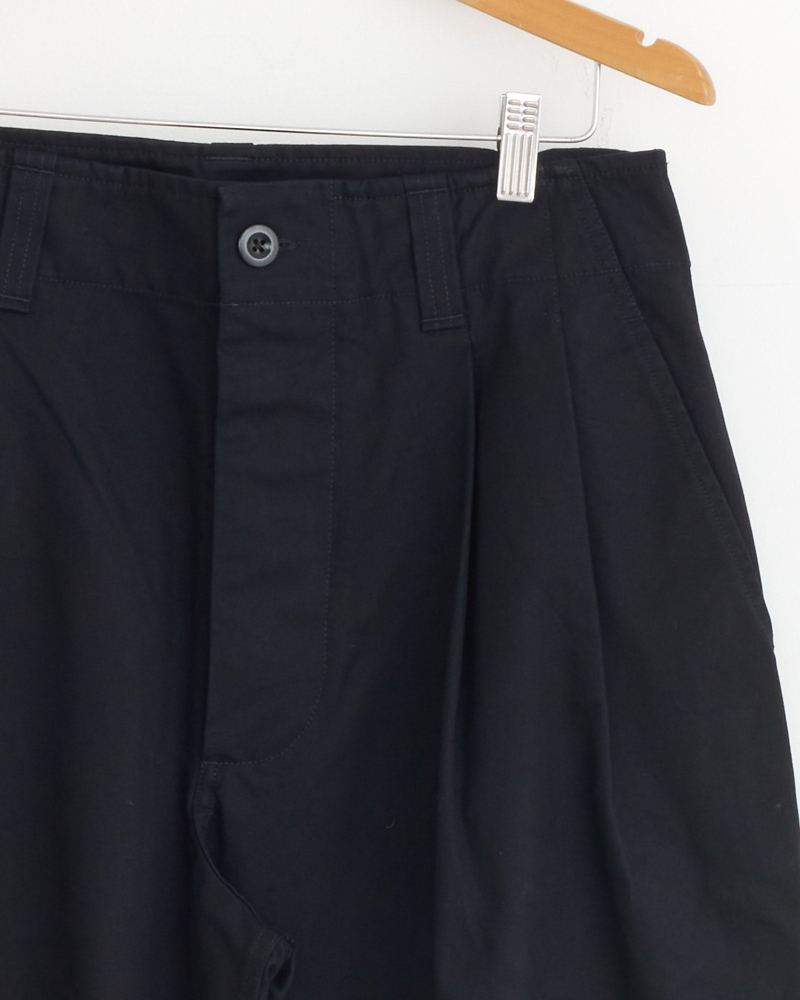 The Stitch Pleat Trouser in Black by MHL by Margaret Howell hangs on a wooden hanger against a white background, shown from waist to upper thigh with button and zipper visible—ideal for checking sizing and fit before purchase.