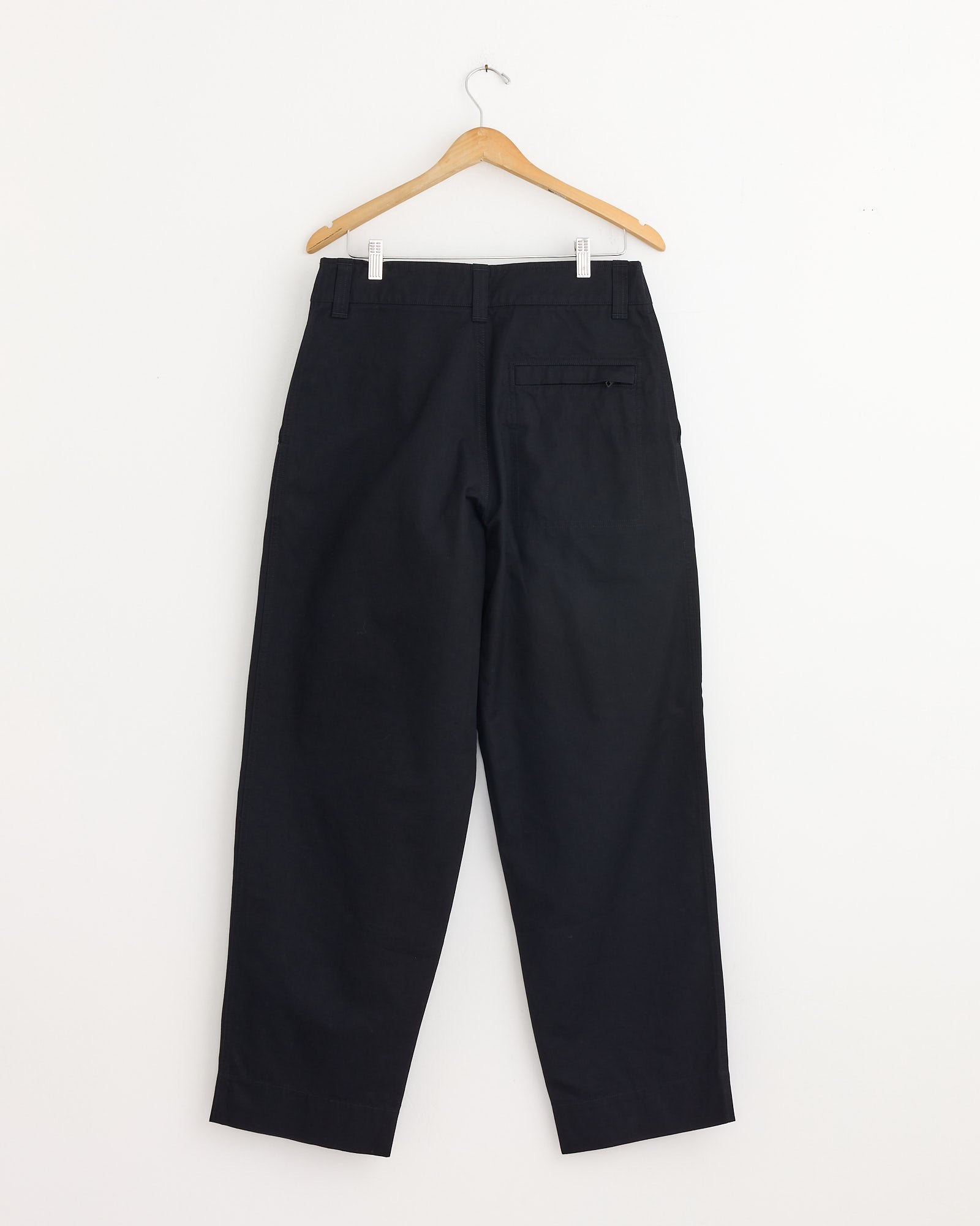 The Stitch Pleat Trouser in Black by MHL by Margaret Howell hangs on a wooden hanger against a white wall, shown from the back with one pocket visible. Questions about fit or sizing? Connect with our online chat support.