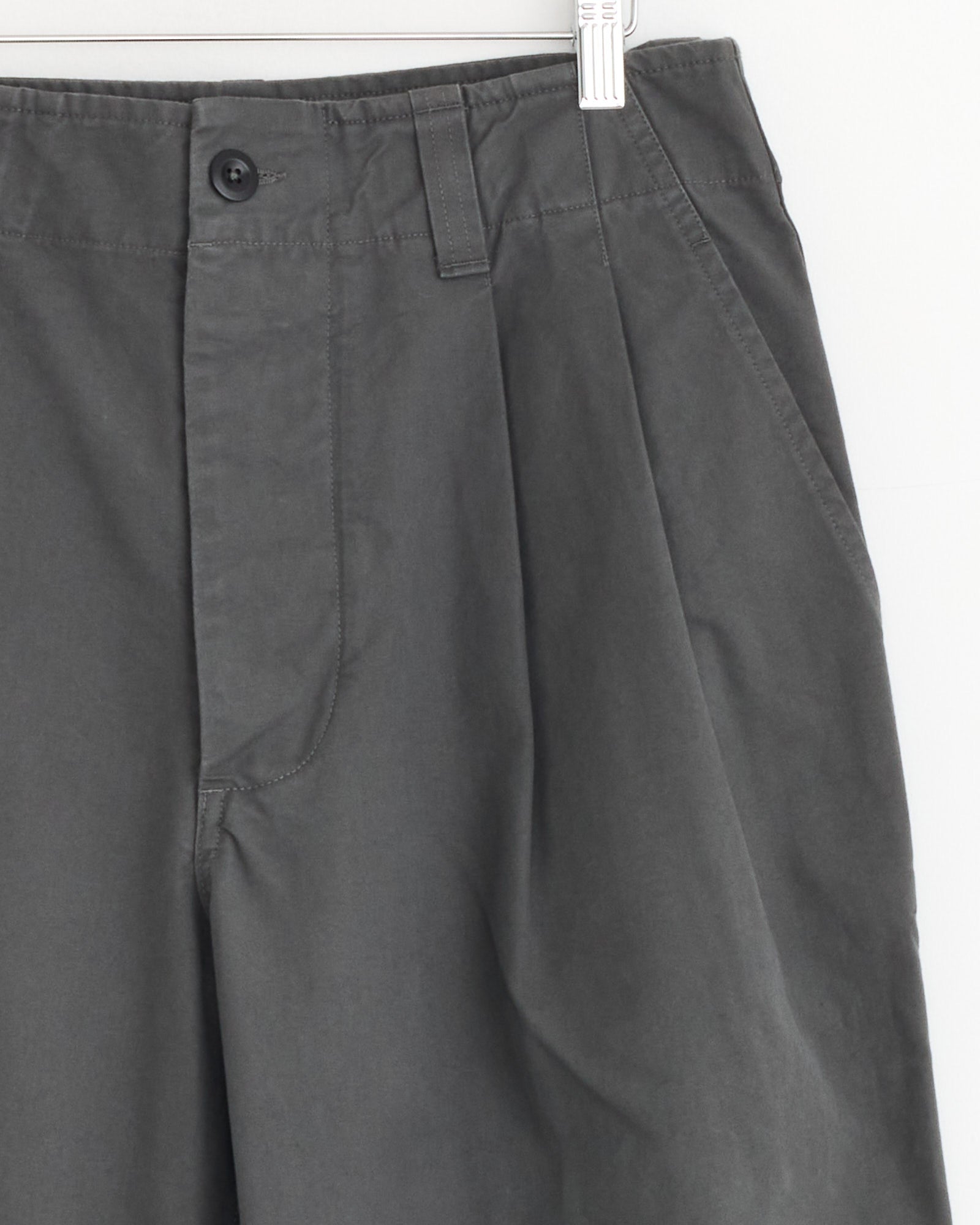 Close-up of the Stitch Pleat Trouser in Charcoal by MHL by Margaret Howell on a white hanger, highlighting waistband, button, belt loop, zipper area, and side pocket against a plain white wall—ideal for viewing fit and sizing details.