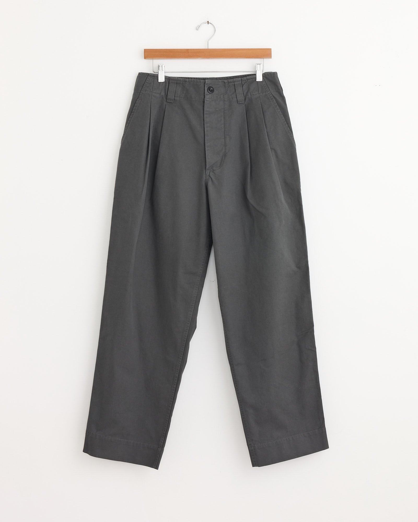 The Stitch Pleat Trouser in Charcoal by MHL by Margaret Howell features a loose, high-waisted fit with pleats, belt loops, and side pockets—ideal for effortless style and comfort. Shown hanging on a wooden hanger against a white background.