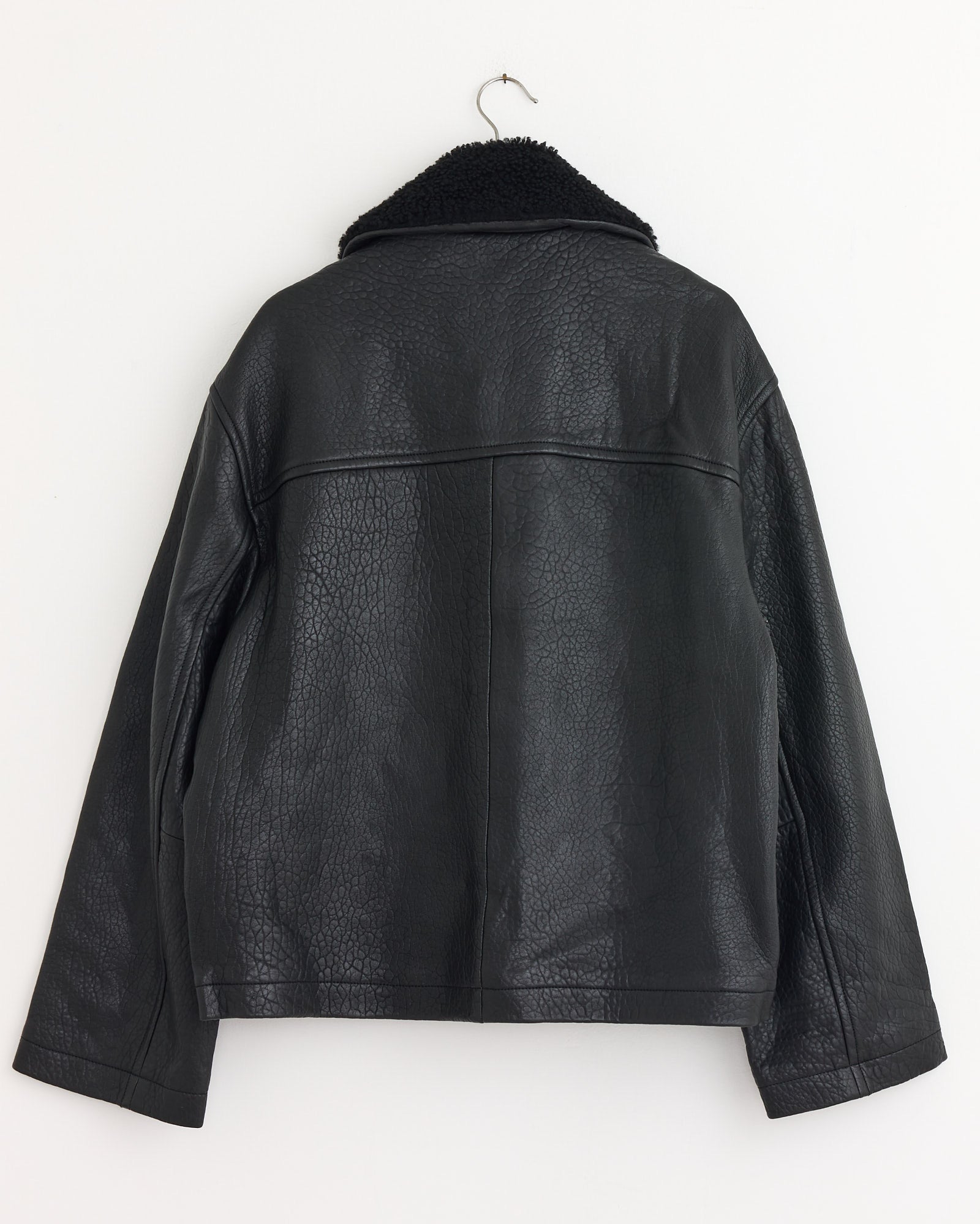 The Studio Nicholson Del Jacket in Black, made from sheepskin leather with a detachable shearling collar, is displayed against a white wall.
