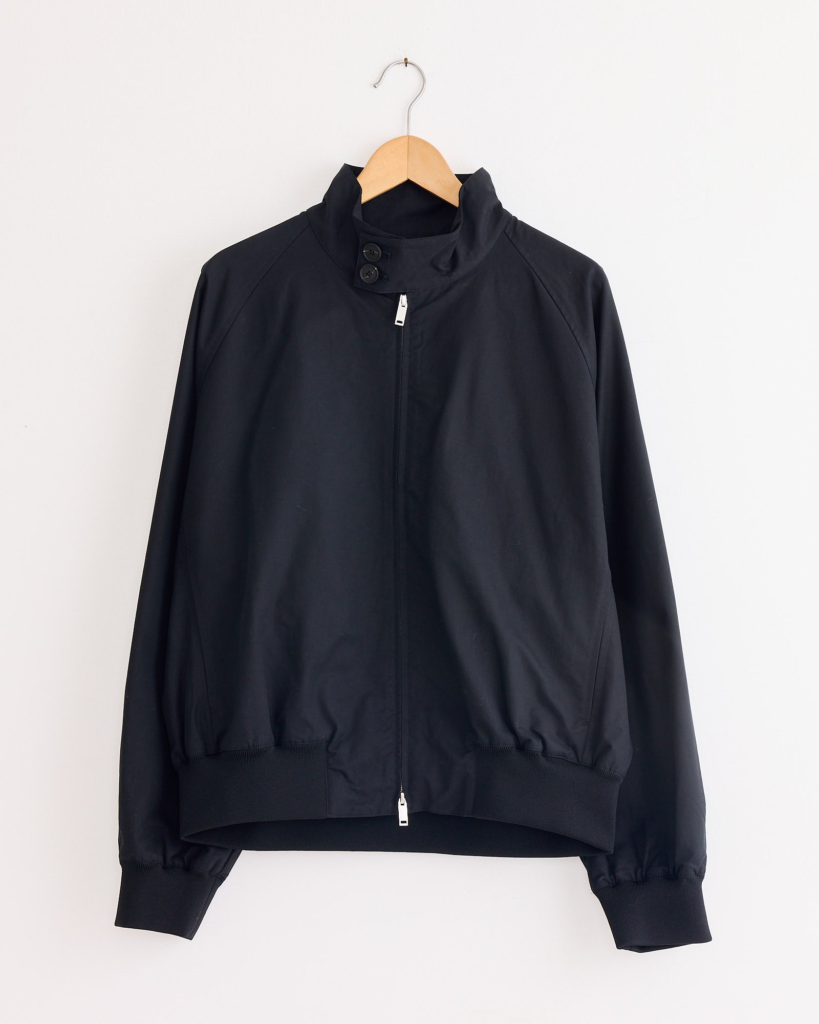 The Studio Nicholson Over Jacket in Oil is a black Harrington jacket with a zip-up front, high buttoned collar, ribbed cuffs and hem, made from showerproof cotton nylon, displayed on a wooden hanger against a white wall.