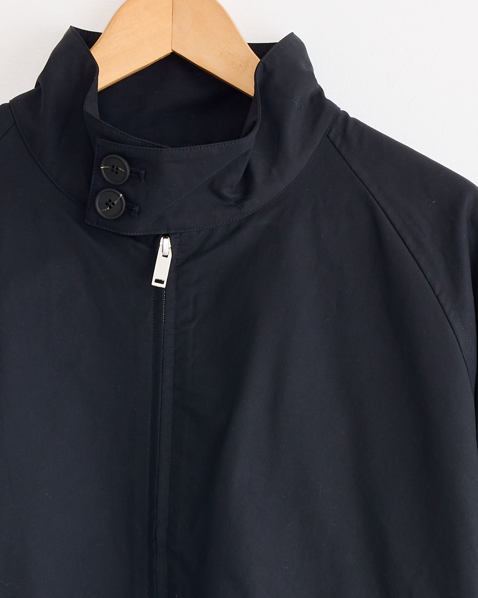 A close-up of the Studio Nicholson Over Jacket in Oil, crafted from showerproof cotton nylon, hangs on a wooden hanger with a high collar, two black buttons, and a partially visible silver zipper.