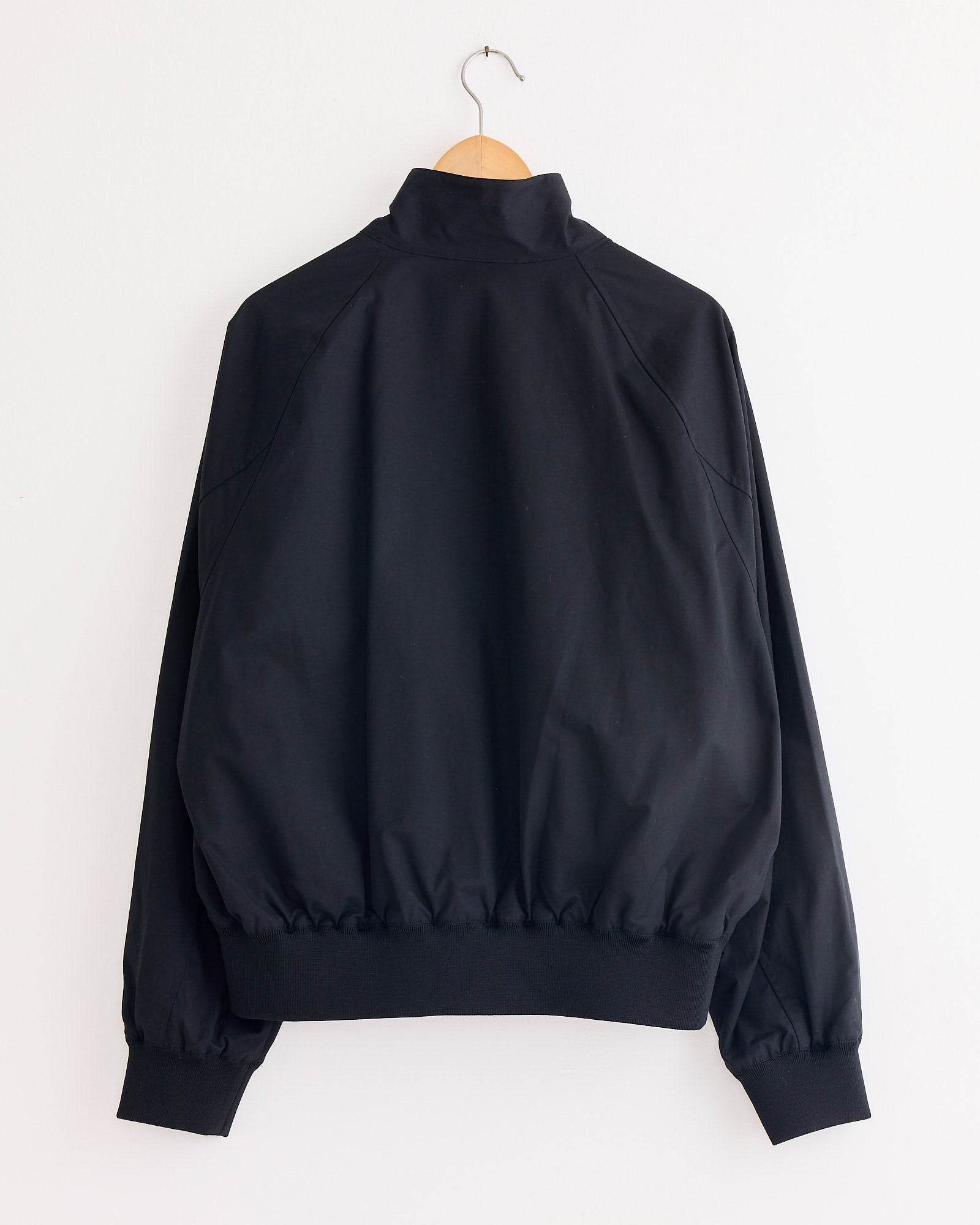 The Studio Nicholson Over Jacket in Oil, a black Harrington-style with long sleeves and high collar, hangs on a wooden hanger against a plain white wall. Made from showerproof cotton-nylon for versatile wear. Shown from the back.