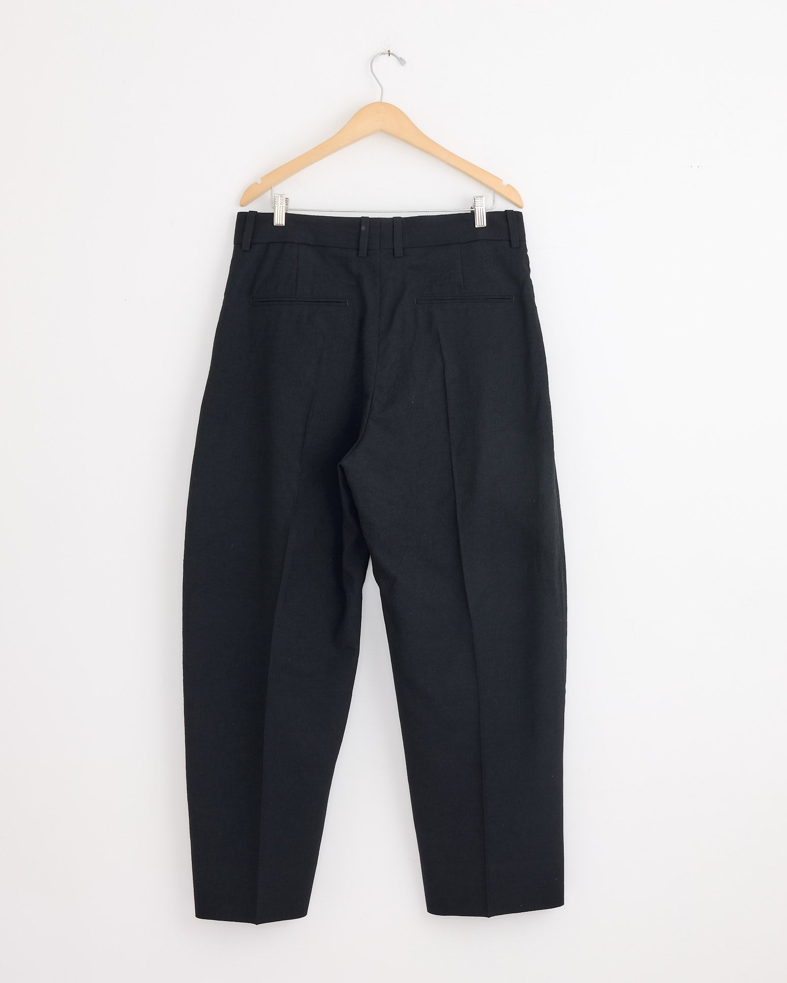 A pair of Studio Nicholson Fume Pants in Black, crafted from Italian-milled virgin wool, hangs on a wooden hanger. They feature belt loops, back pockets, and a relaxed fit with a tapered leg.
