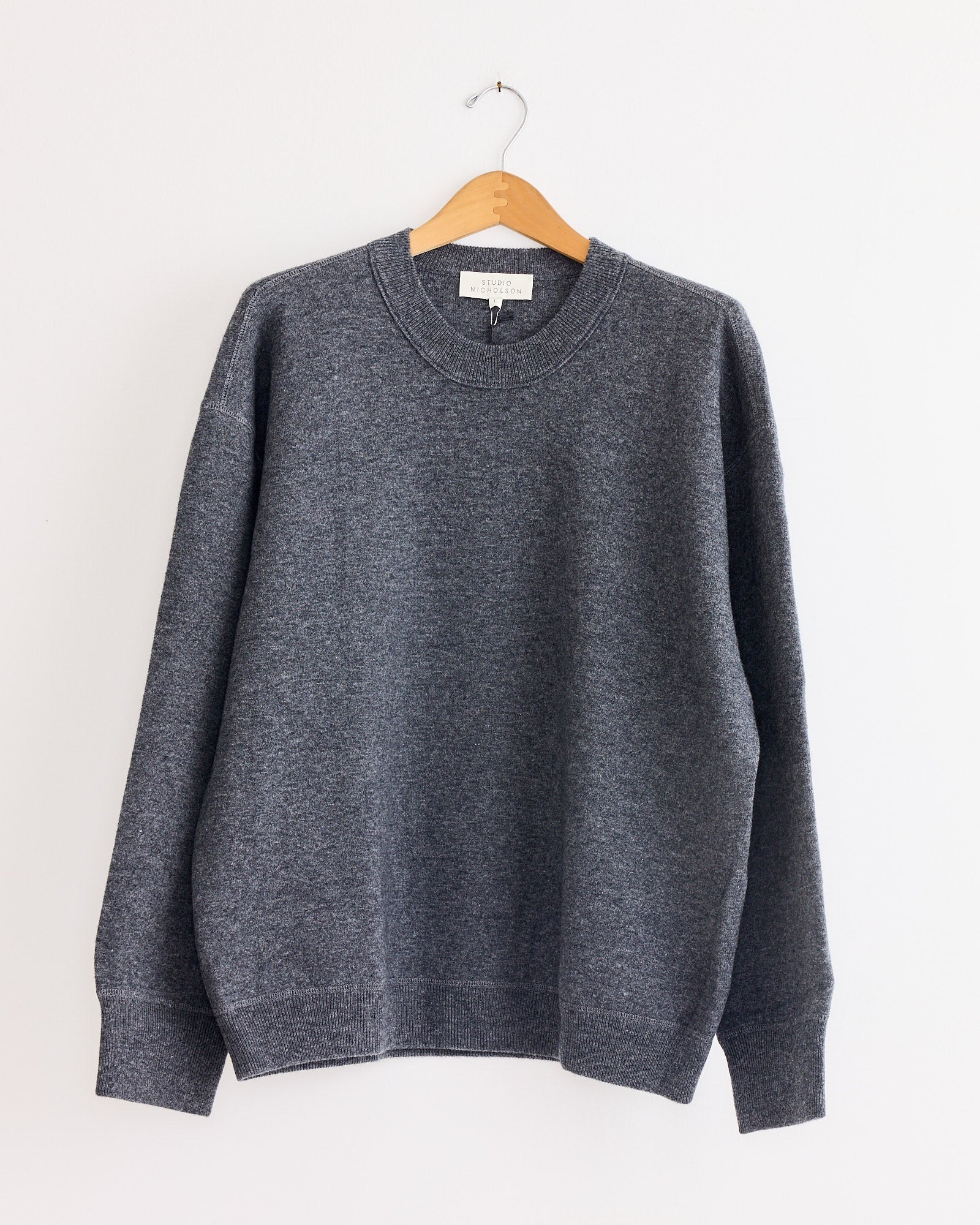 The Studio Nicholson Moray Sweatshirt in Charcoal Marl, crafted from double cashmere, hangs on a wooden hanger against a white wall.