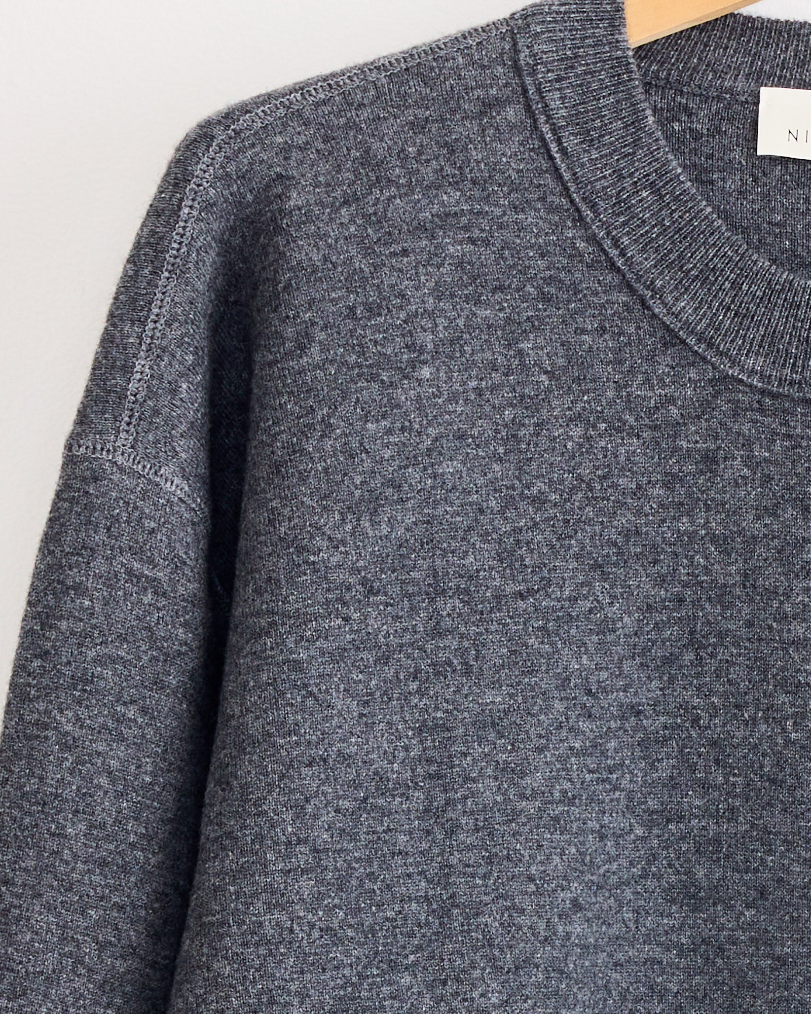 A close-up of the Studio Nicholson Moray Sweatshirt in Charcoal Marl, made from double cashmere, hangs on a wooden hanger, showcasing its ribbed collar and shoulder seam against a light background.