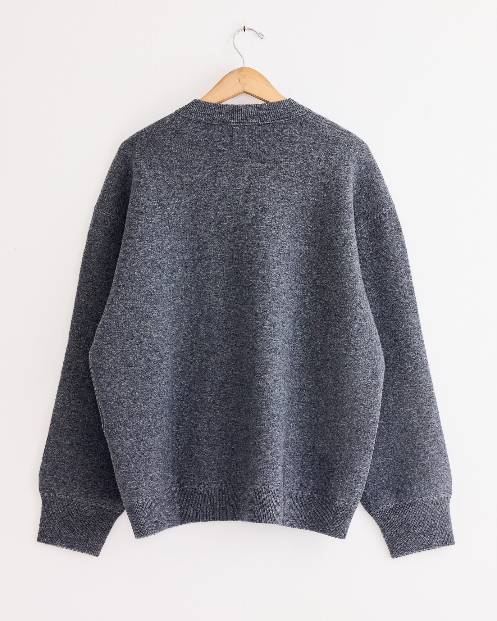 The Studio Nicholson Moray Sweatshirt in Charcoal Marl, featuring long sleeves and double cashmere fabric, is displayed from the back on a wooden hanger against a white wall.