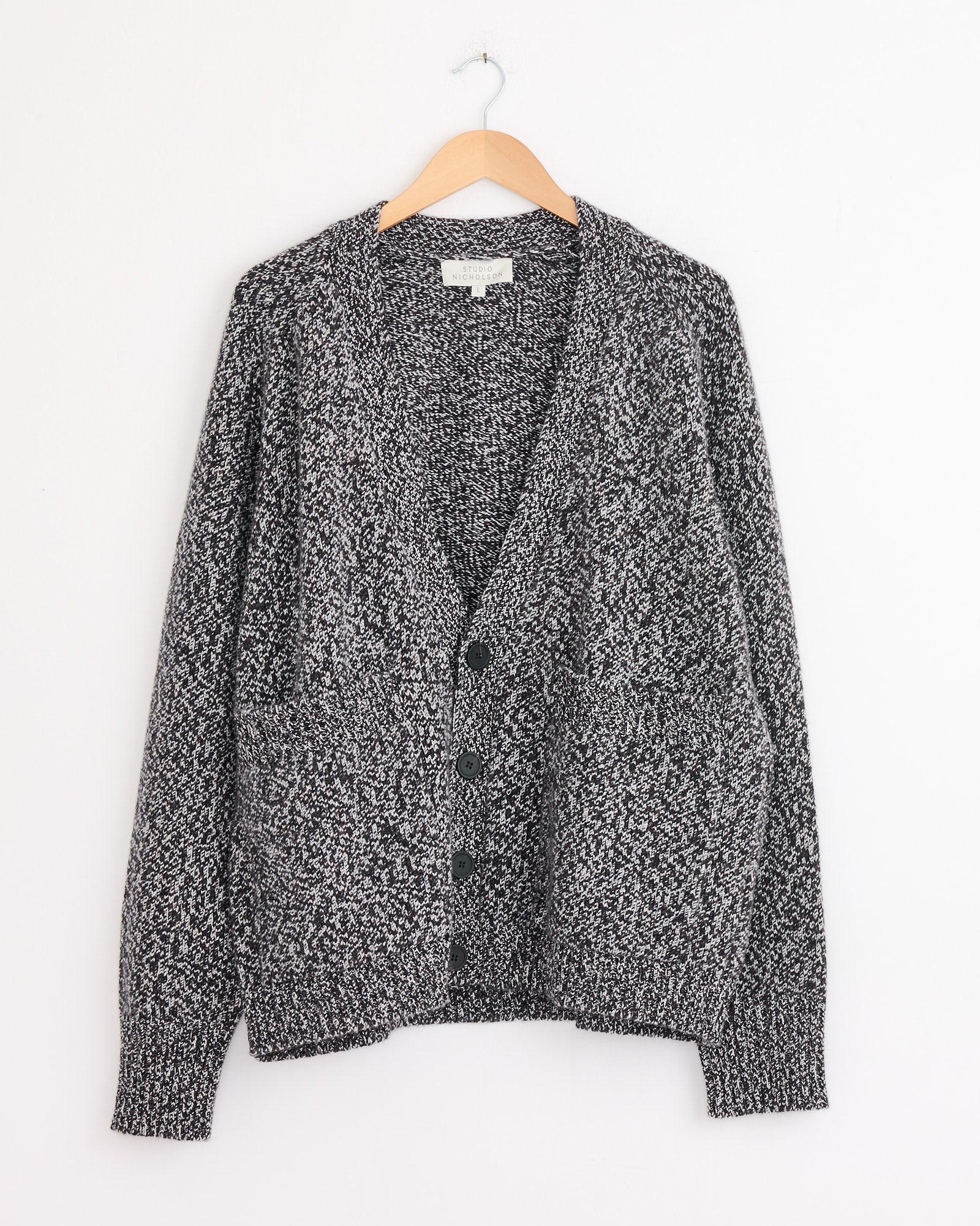 A Studio Nicholson Sen Cardigan in navy mouline heavyweight knit with long sleeves, front pockets, and black buttons, hangs on a wooden hanger against a white wall.