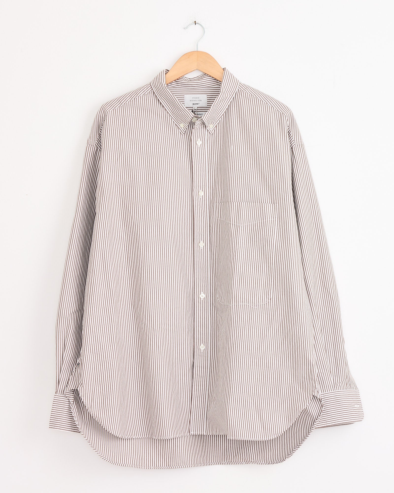 The Studio Nicholson x BEAMS PLUS Lisa Shirt in Brown/White Stripe, a long-sleeve oxford shirt with thin vertical stripes, a collar, chest pocket, and front white buttons, hangs on a wooden hanger against a plain white wall.