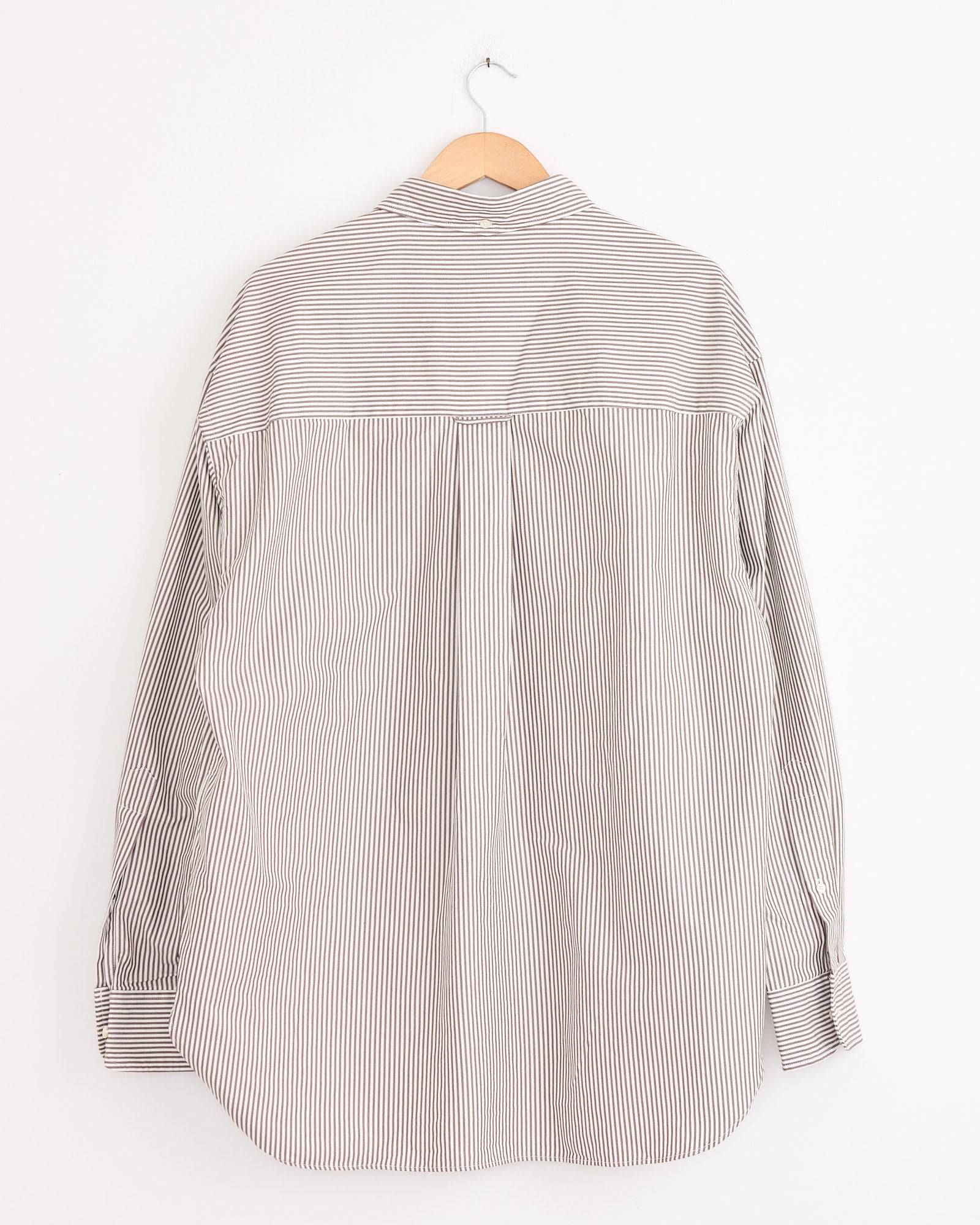 The Studio Nicholson x BEAMS PLUS Lisa Shirt in Brown/White Stripe—long-sleeved, made in Japan with thin brown and white vertical stripes—is shown from the back on a wooden hanger against a white wall.