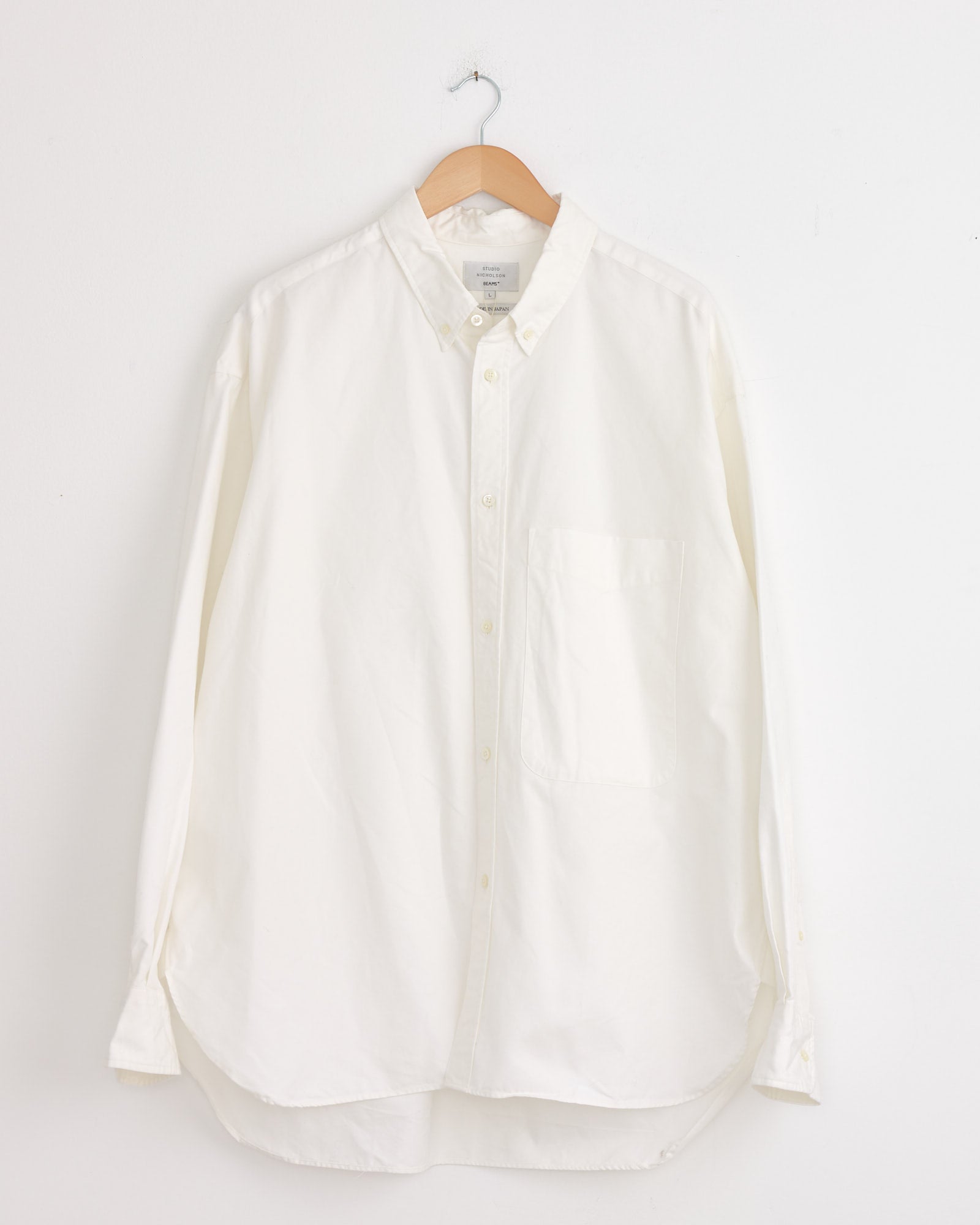 The Studio Nicholson x BEAMS PLUS Lisa Shirt in White, crafted in Japan, hangs on a wooden hanger against a white wall. This Oxford cloth shirt features a front chest pocket and a pointed collar for timeless style.