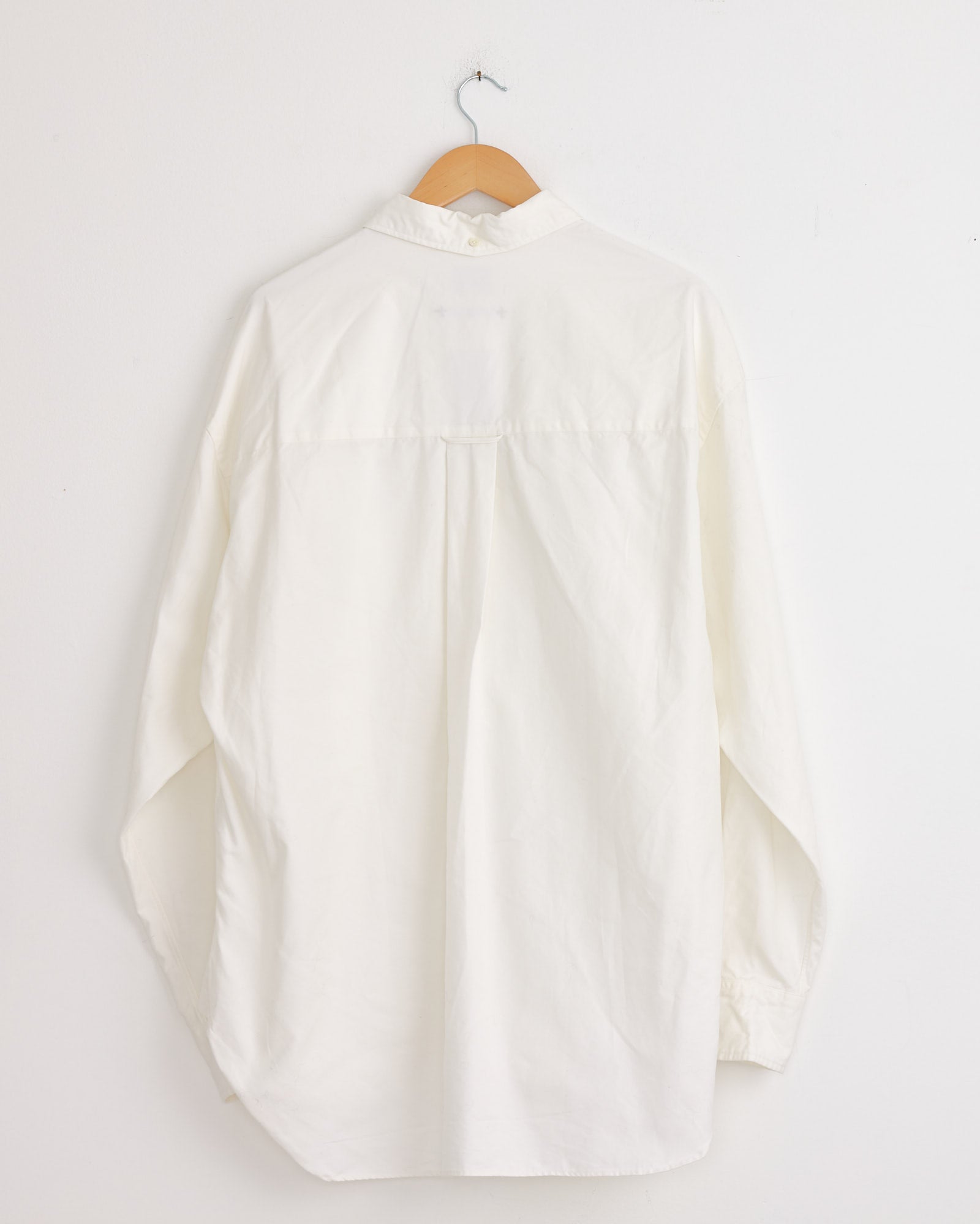The Studio Nicholson x BEAMS PLUS Lisa Shirt in White, a long-sleeved Oxford shirt, is displayed from the back on a wooden hanger against a plain white wall.