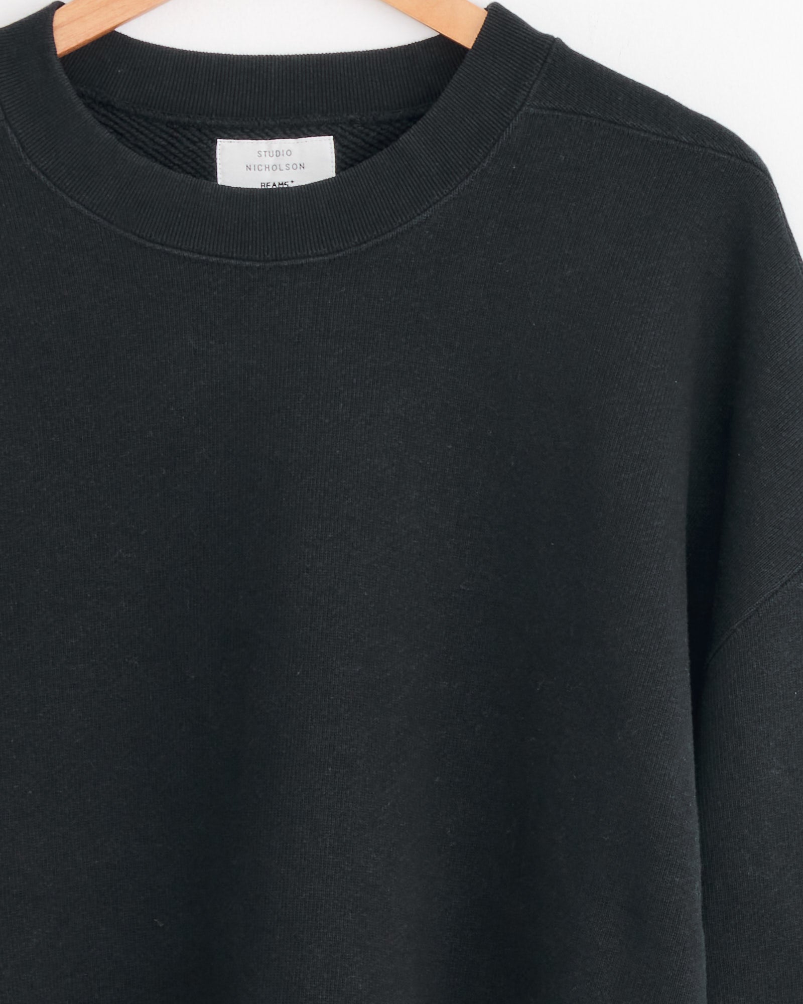 A close-up of the Studio Nicholson x BEAMS PLUS Holly Crewneck in Darkest Navy, made in Japan, hangs on a wooden hanger, revealing the Studio Nicholson label inside the collar.