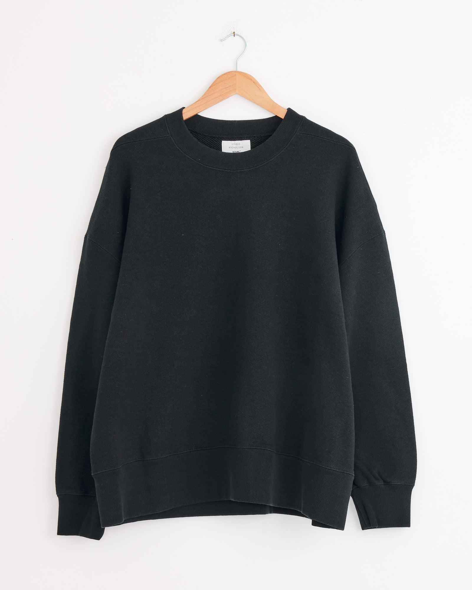 The Studio Nicholson x BEAMS PLUS Holly Crewneck in Darkest Navy, a long-sleeve sweatshirt, hangs on a wooden hanger against a white wall.