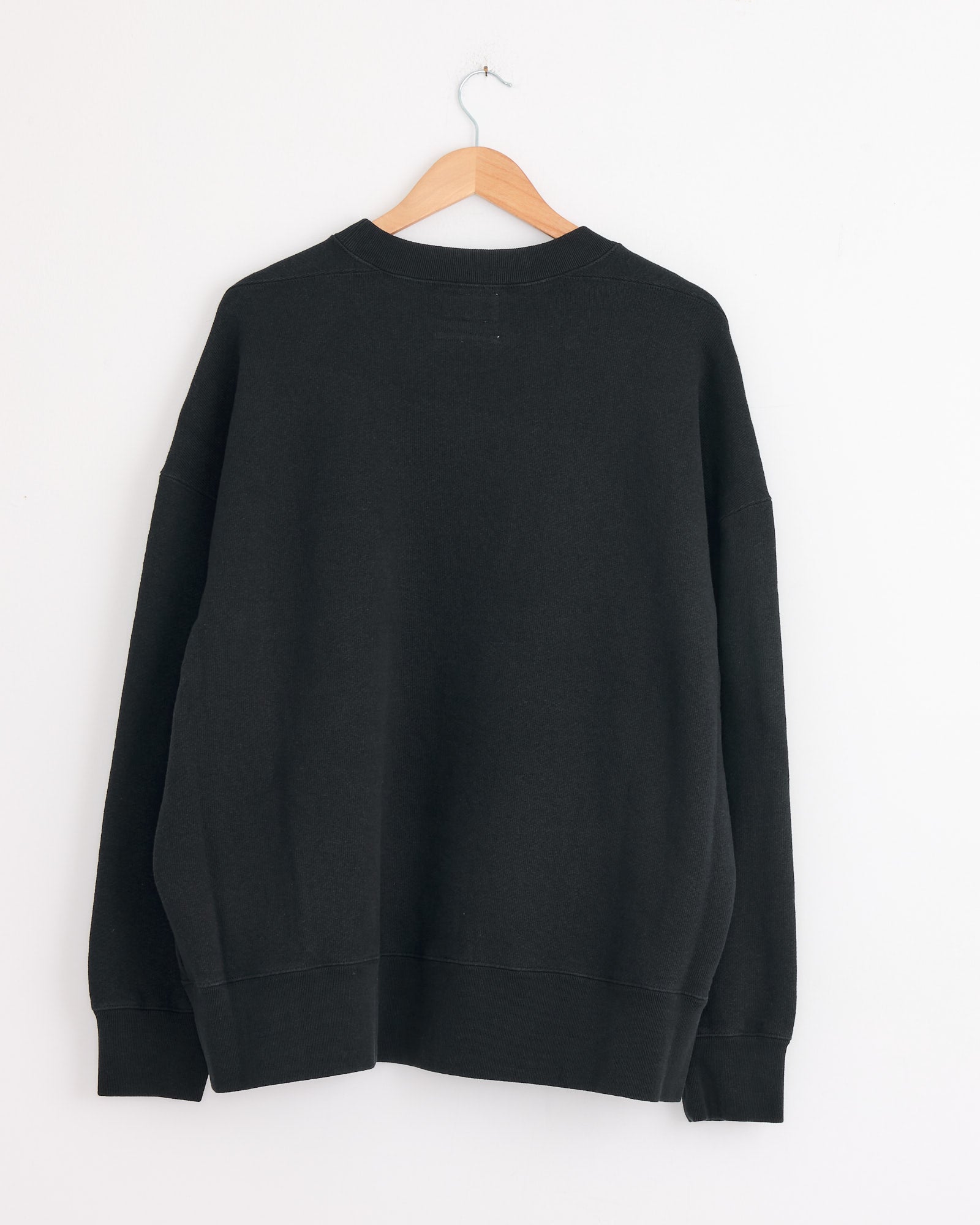 The Studio Nicholson x BEAMS PLUS Holly Crewneck in Darkest Navy by Studio Nicholson, made in Japan, is displayed from the back on a wooden hanger against a white wall.