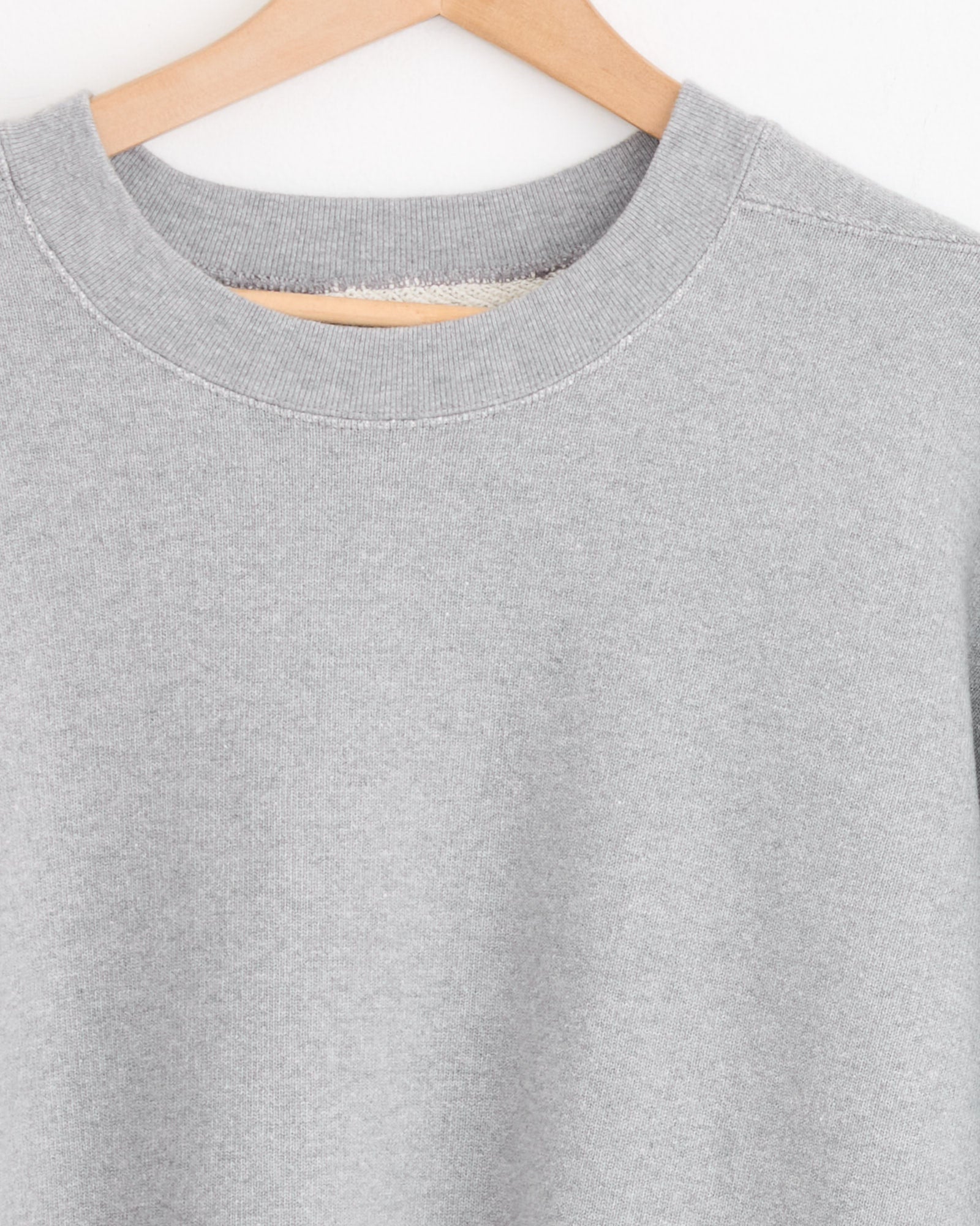 The Studio Nicholson x BEAMS PLUS Holly Crewneck in Grey Marl hangs on a wooden hanger against a white background, with the image focusing on the collar and shoulders.