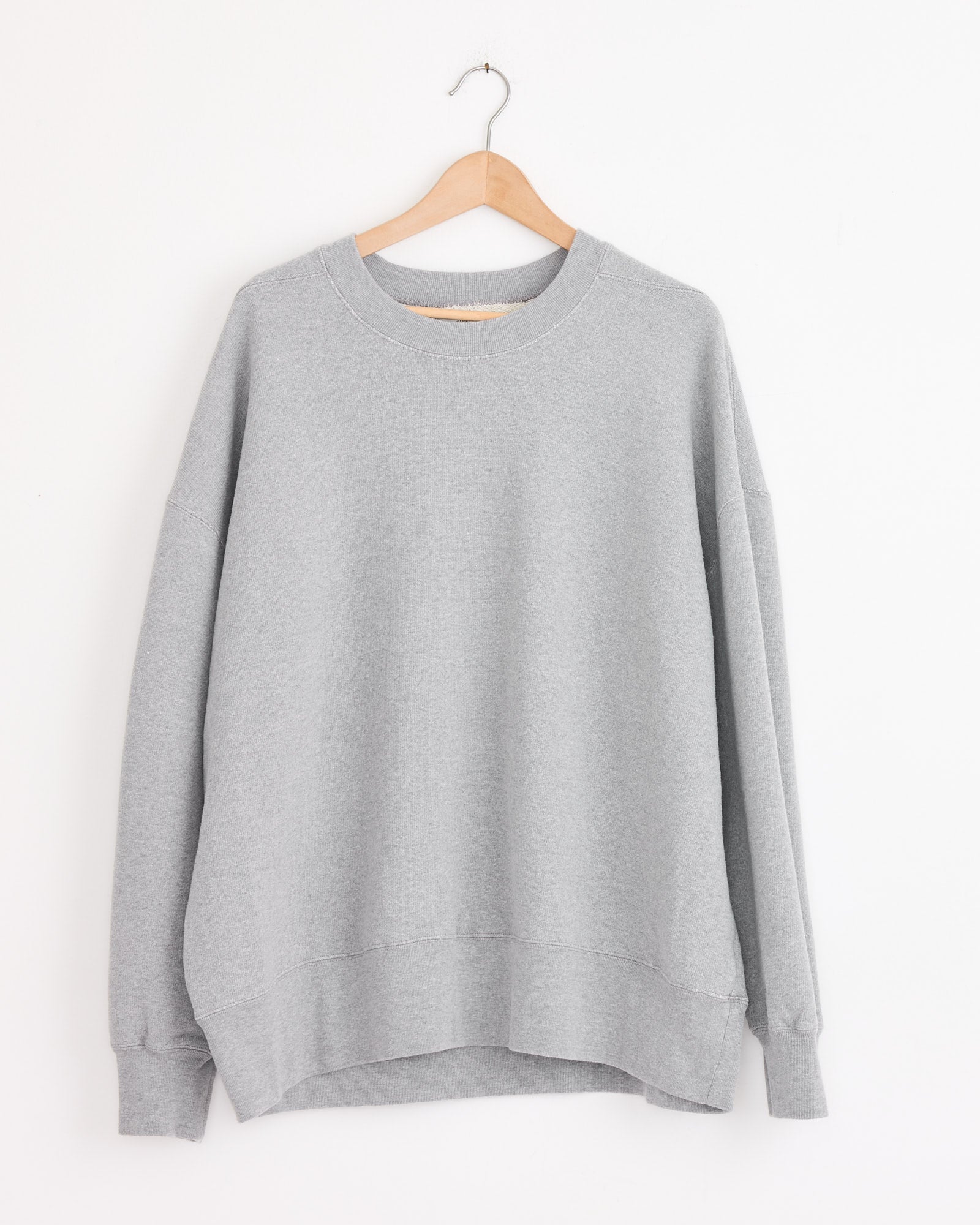 The Studio Nicholson x BEAMS PLUS Holly Crewneck in Grey Marl, a light gray long-sleeve sweatshirt, hangs on a wooden hanger against a white wall.
