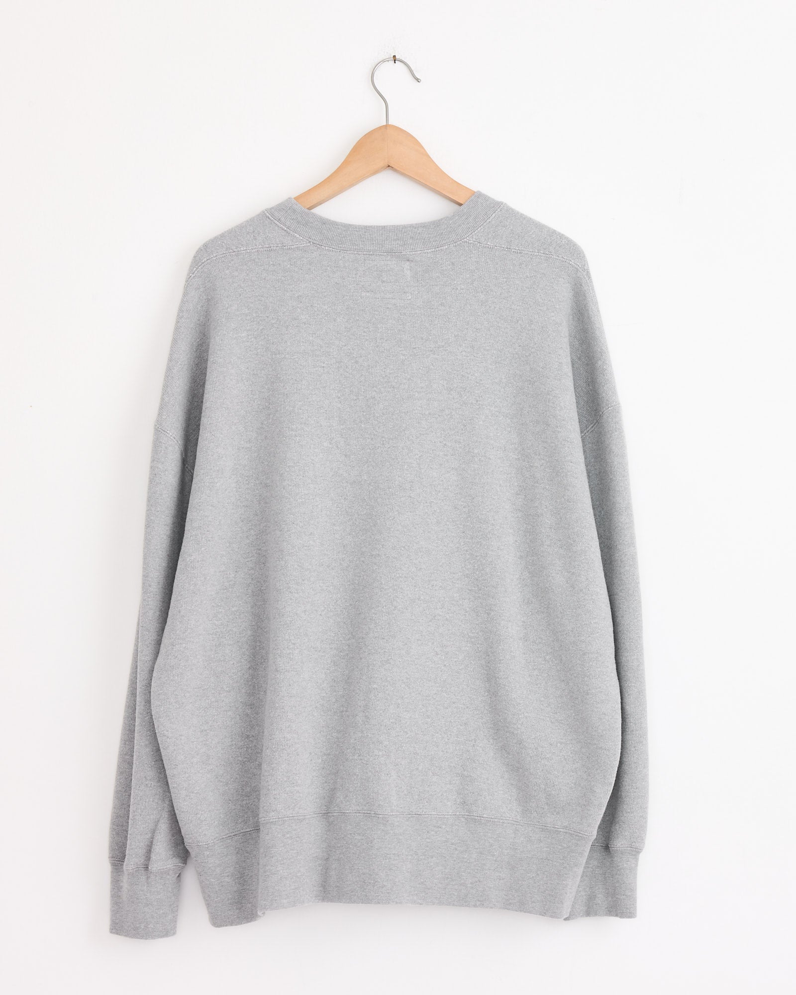 The Studio Nicholson x BEAMS PLUS Holly Crewneck in Grey Marl hangs on a wooden hanger against a white wall, displayed with the back facing forward.