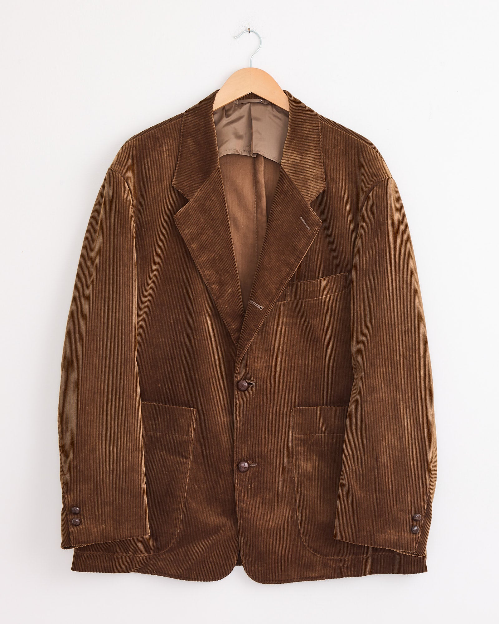 The Studio Nicholson x BEAMS PLUS Paul Coat in Toffee, inspired by the classic Paul Coat, is a brown corduroy blazer with notch lapels, three front pockets, and buttoned cuffs, hanging on a wooden hanger against a white wall.