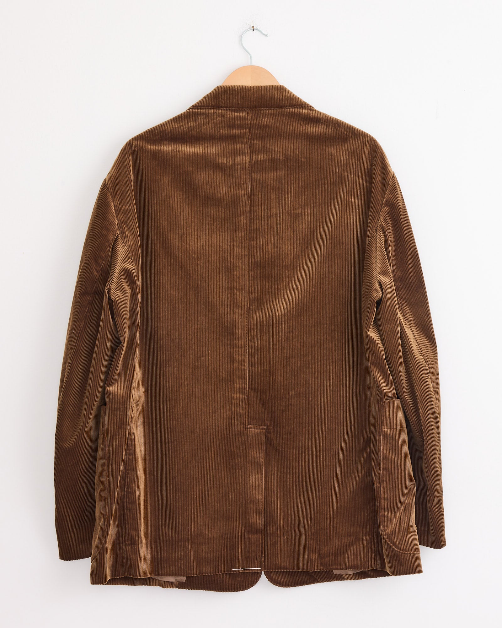 The Studio Nicholson x BEAMS PLUS Paul Coat in Toffee, crafted in Japan, hangs on a wooden hanger against a white wall, displayed from the back to showcase its distinctive texture and stitching.