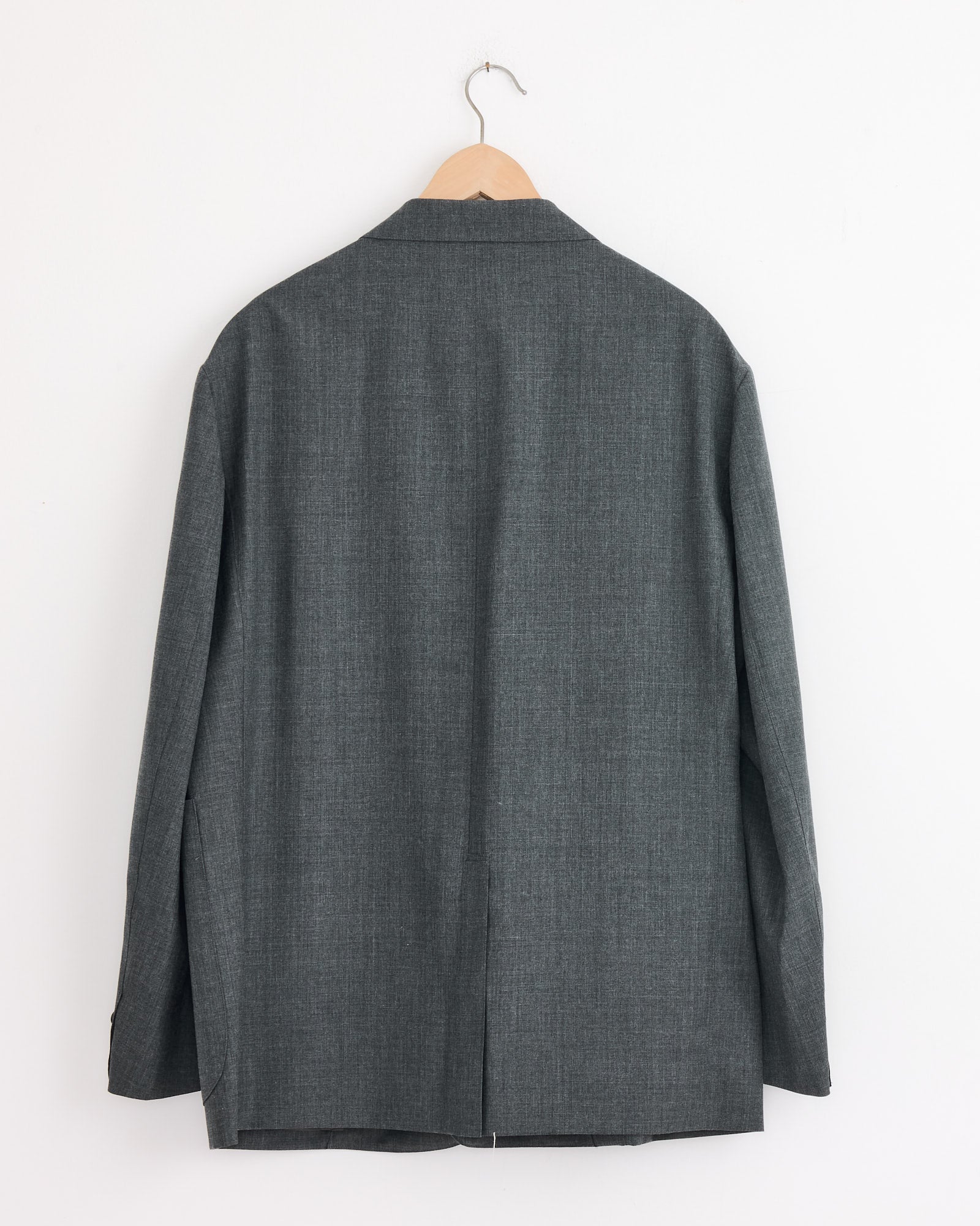 The Studio Nicholson x BEAMS PLUS Paul Coat in Charcoal Marl, made in Japan, is displayed from the back on a wooden hanger against a white wall. The wool blazer’s fabric looks smooth with subtle texture.