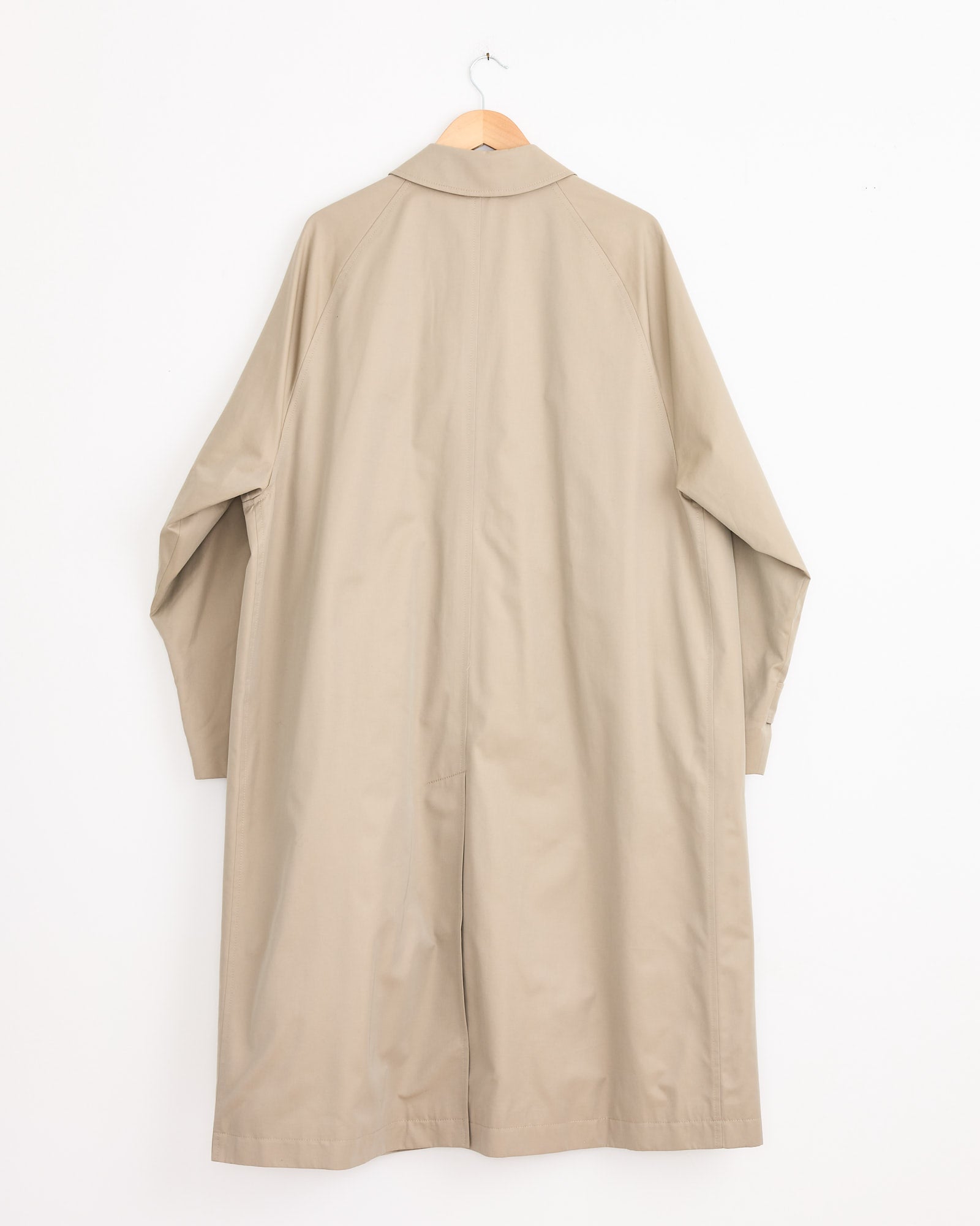 The Studio Nicholson x BEAMS PLUS Peter Raincoat in Tan, made from water-repellent gabardine, hangs on a wooden hanger against a plain white wall, shown from the back.