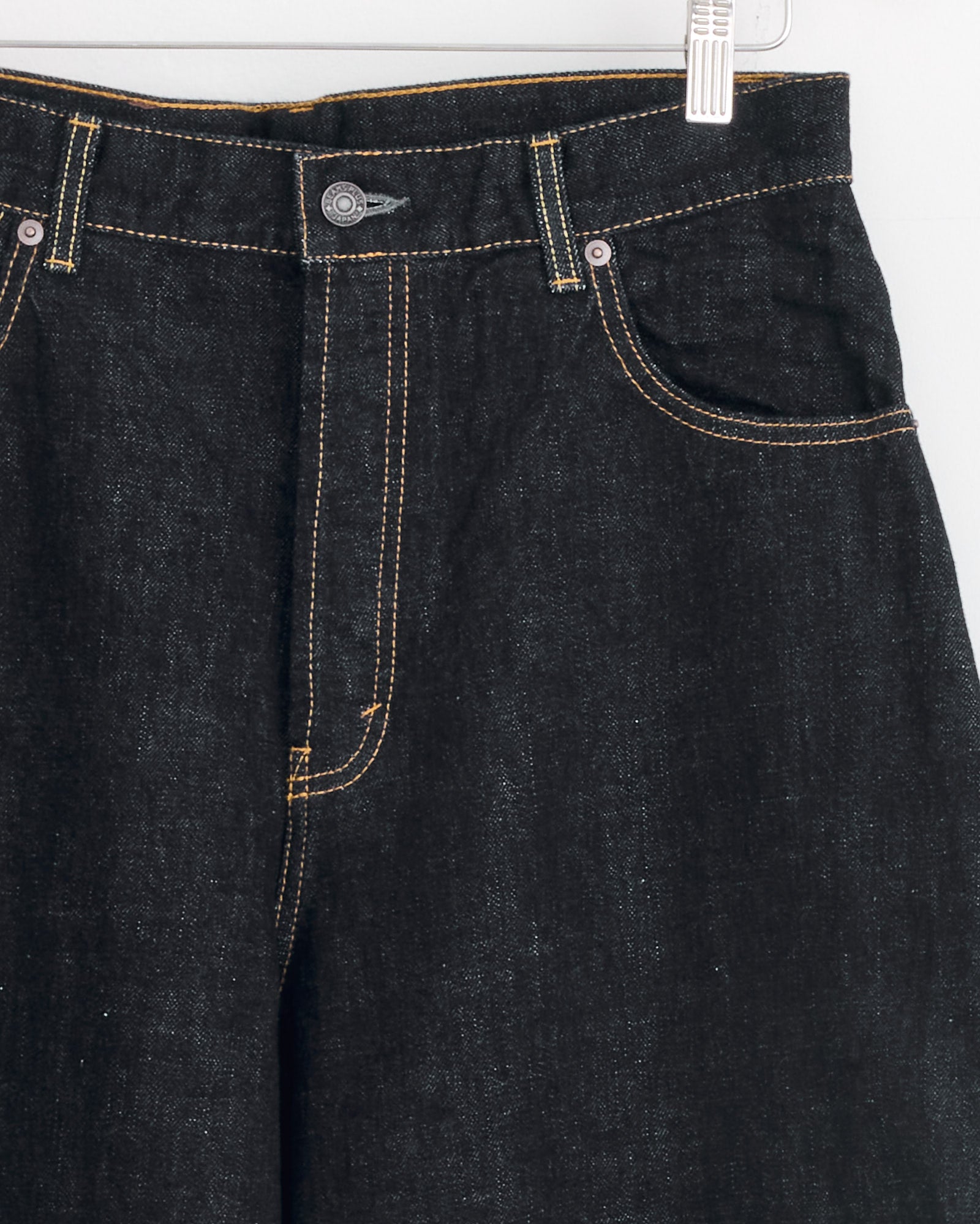 Close-up of Studio Nicholson x BEAMS PLUS Holly Jean in Indigo—Japanese selvedge denim with yellow stitching, metal button, and front pockets—hanging on a white hanger against a light background.