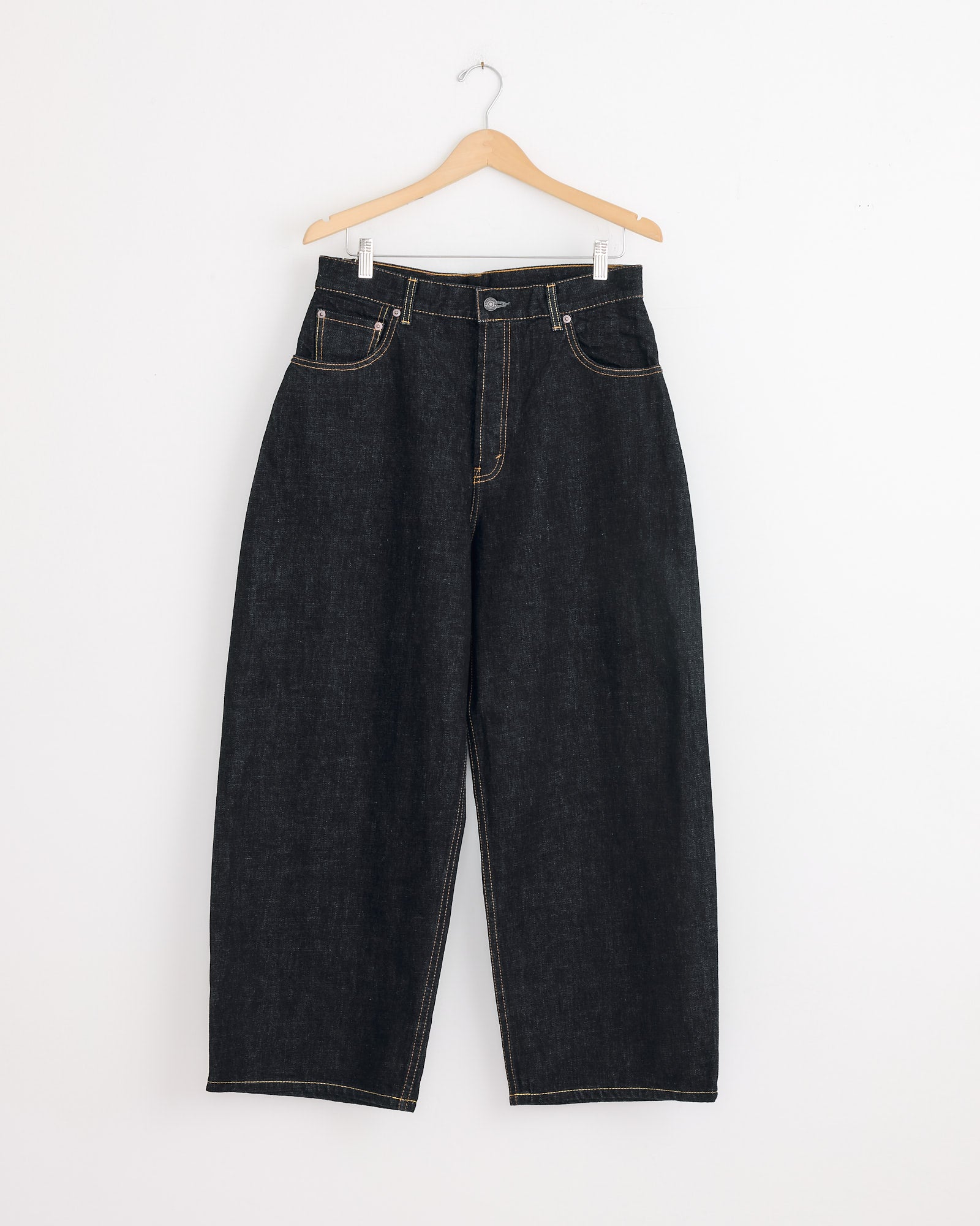 The Studio Nicholson x BEAMS PLUS Holly Jean in Indigo features a high-waisted, wide-leg balloon silhouette in Japanese selvedge denim with contrast stitching, shown on a wooden hanger against a plain white wall.