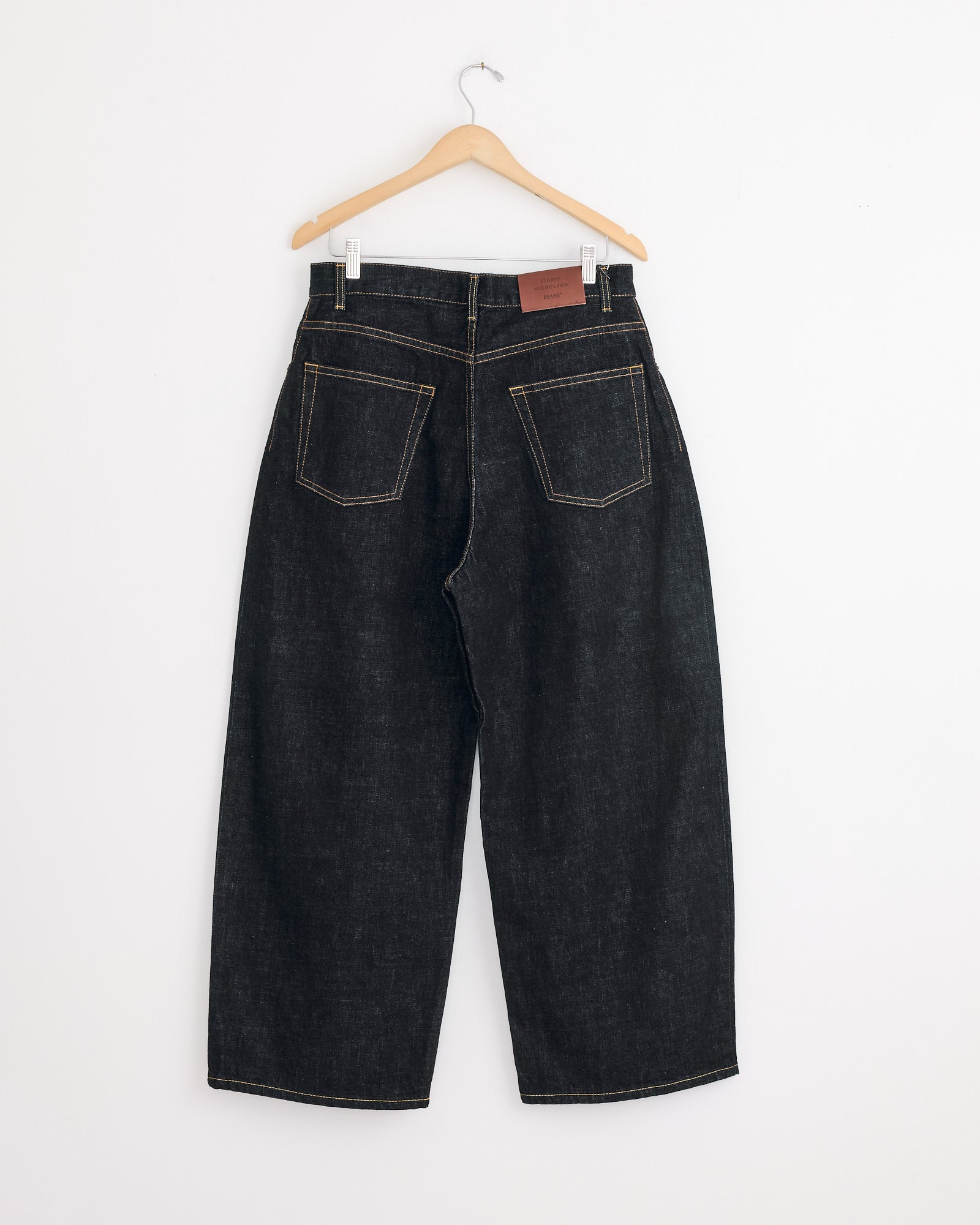 The Studio Nicholson x BEAMS PLUS Holly Jean in Indigo, featuring balloon legs and large back pockets, crafted from Japanese selvedge denim, hangs on a wooden hanger against a plain white wall.