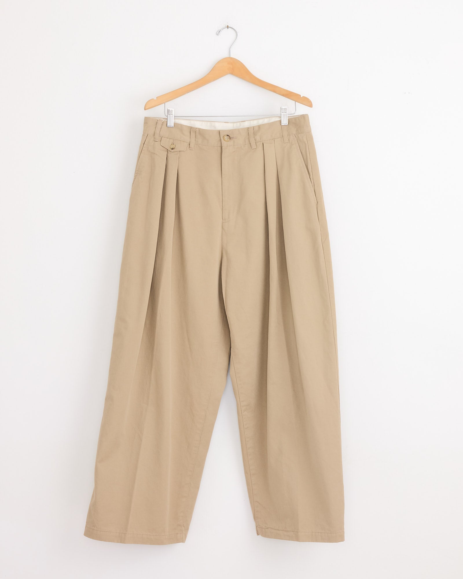 The Studio Nicholson x BEAMS PLUS Alvy Pant in Tan features a loose, high-waisted fit with pleats, belt loops, and side pockets in high-density cotton twill, displayed on a wooden hanger against a white background.