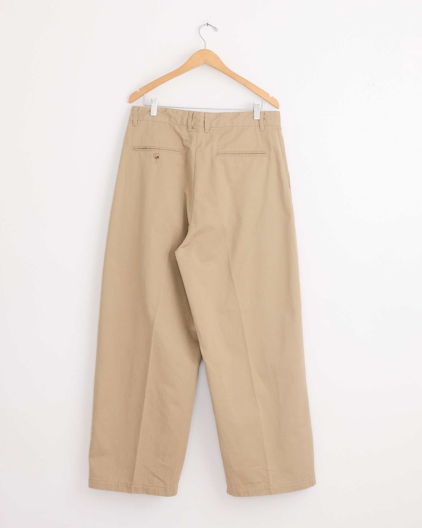 The Studio Nicholson x BEAMS PLUS Alvy Pant in Tan, made from high-density cotton twill, hangs on a wooden hanger against a white wall, shown from the back with two welt pockets—one has a button.