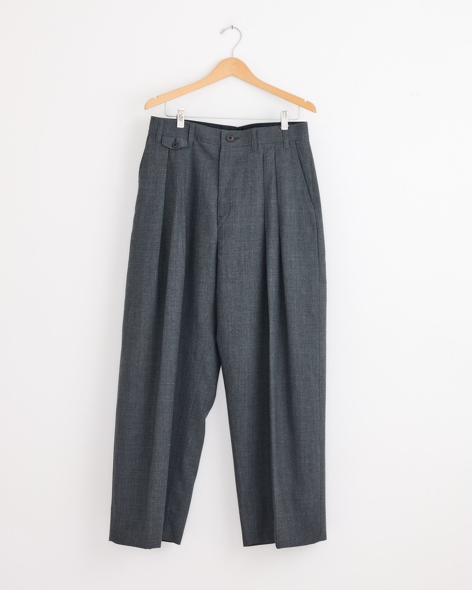The Studio Nicholson x BEAMS PLUS Alvy Pant in Charcoal Marl, inspired by 1970s menswear, hangs on a wooden hanger against a plain white wall.