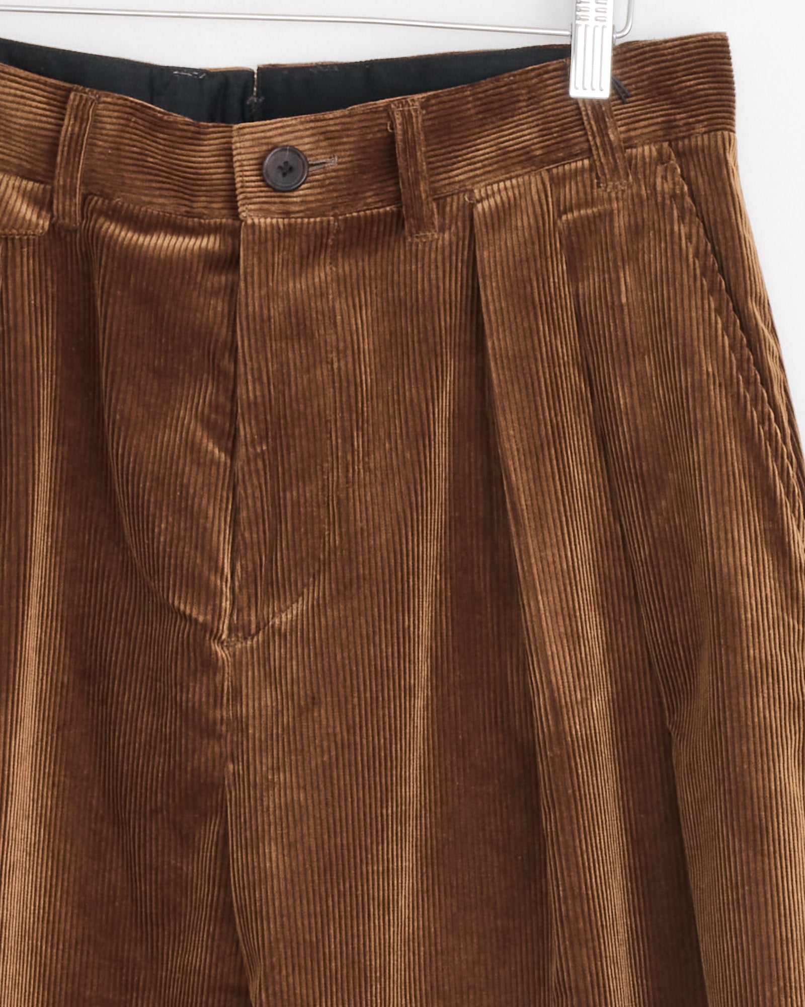 Close-up of Studio Nicholson x BEAMS PLUS Alvy Pant in Toffee, featuring vertical corduroy ridges, pleats, black button, and belt loops, hanging on a hanger against a light background.