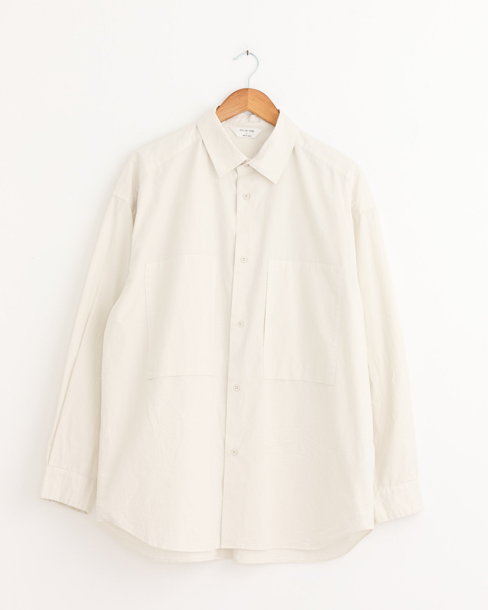 The Still By Hand Garment Dye Over Shirt in Ecru, crafted in Japan from soft peach skin cotton, features a collar, two large front pockets, and long sleeves. It hangs on a wooden hanger against a white background.