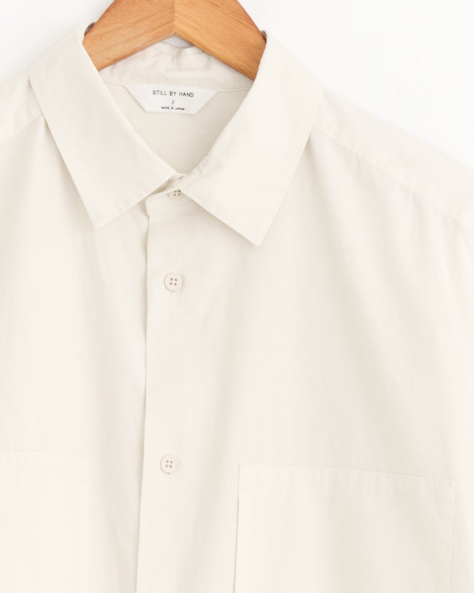 A close-up of the Still By Hand Garment Dye Over Shirt in Ecru, made in Japan and shown on a wooden hanger. It has a classic collar, two chest pockets, and the Still By Hand label inside.