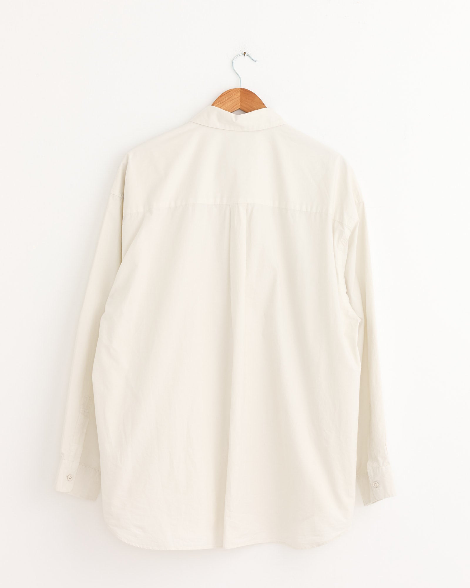 The Still By Hand Garment Dye Over Shirt in Ecru, a long-sleeved, Japanese-made piece, hangs on a wooden hanger against a plain white wall, displayed from the back.