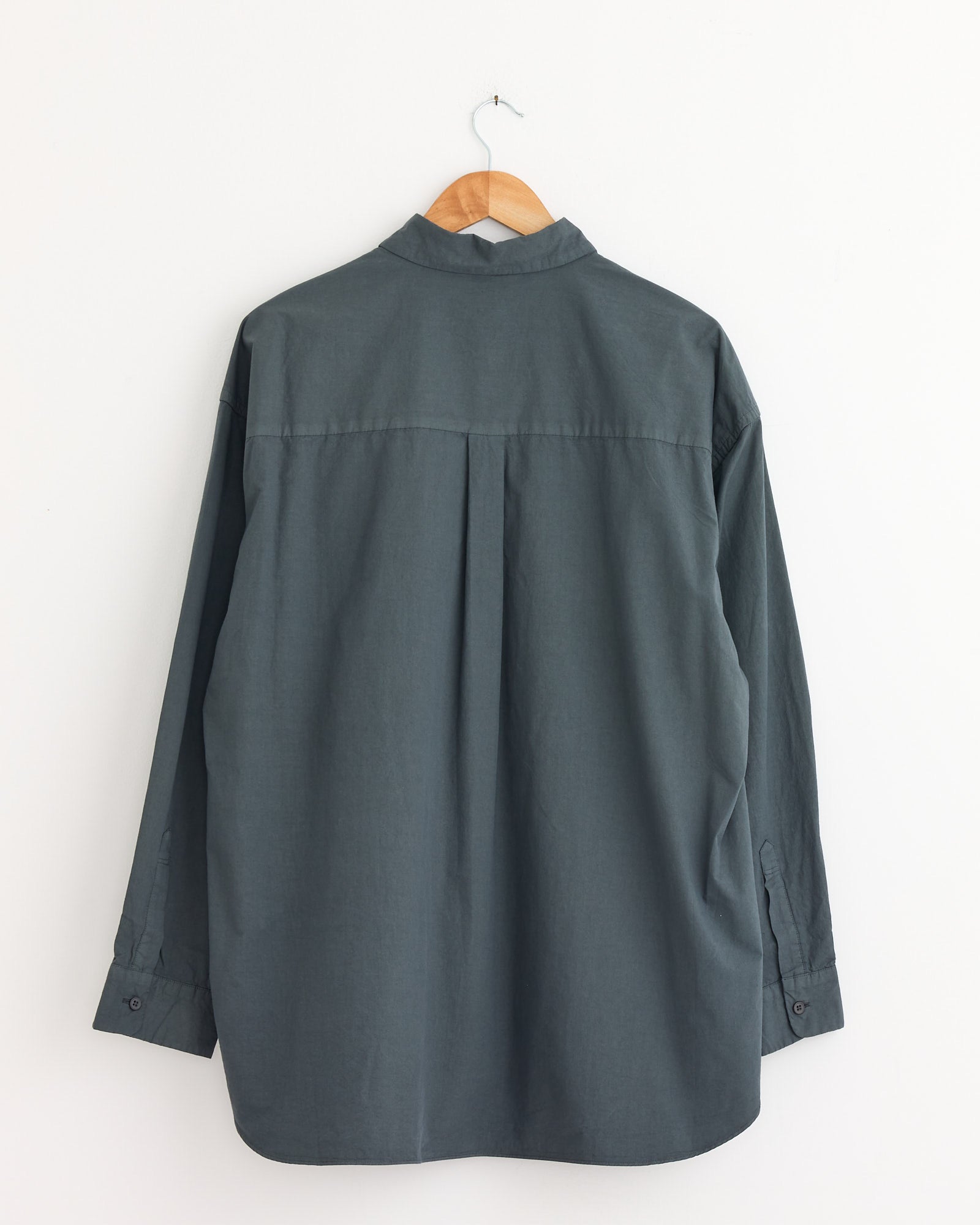 The Garment Dye Over Shirt in Blue Grey by Still By Hand hangs on a wooden hanger against a plain white wall, shown from the back.