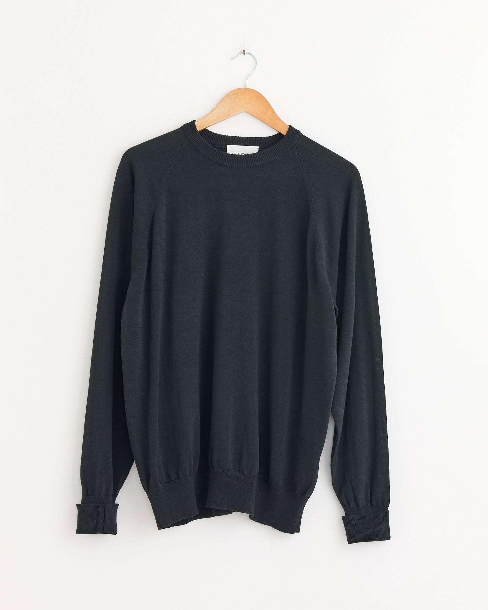 The Cotton Silk Pullover in Green Charcoal by Still By Hand, crafted in Japan, hangs on a wooden hanger against a white wall, highlighting its minimalist style and lightweight design.