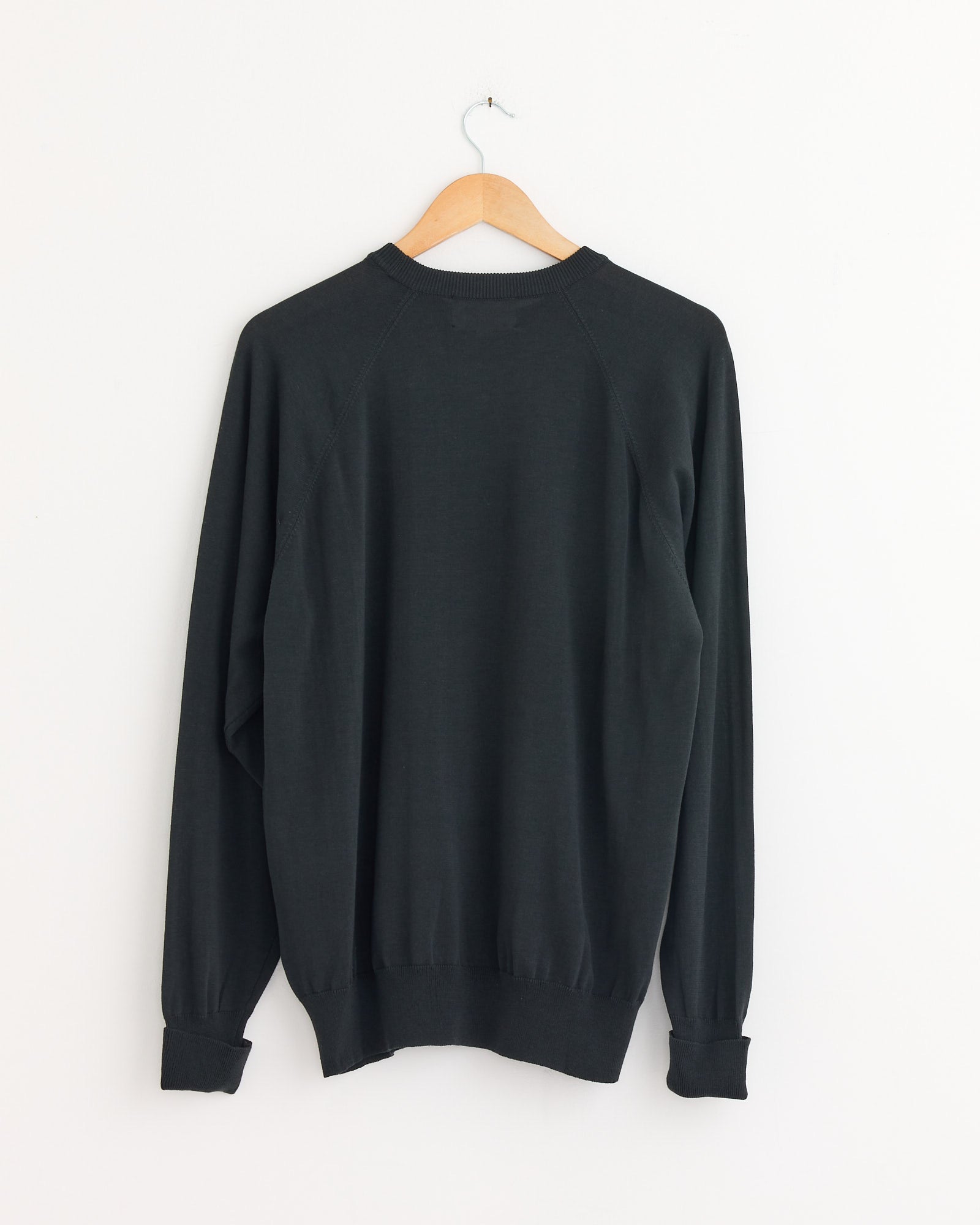 A Cotton Silk Pullover in Green Charcoal by Still By Hand hangs on a wooden hanger against a white wall, displayed from the back.