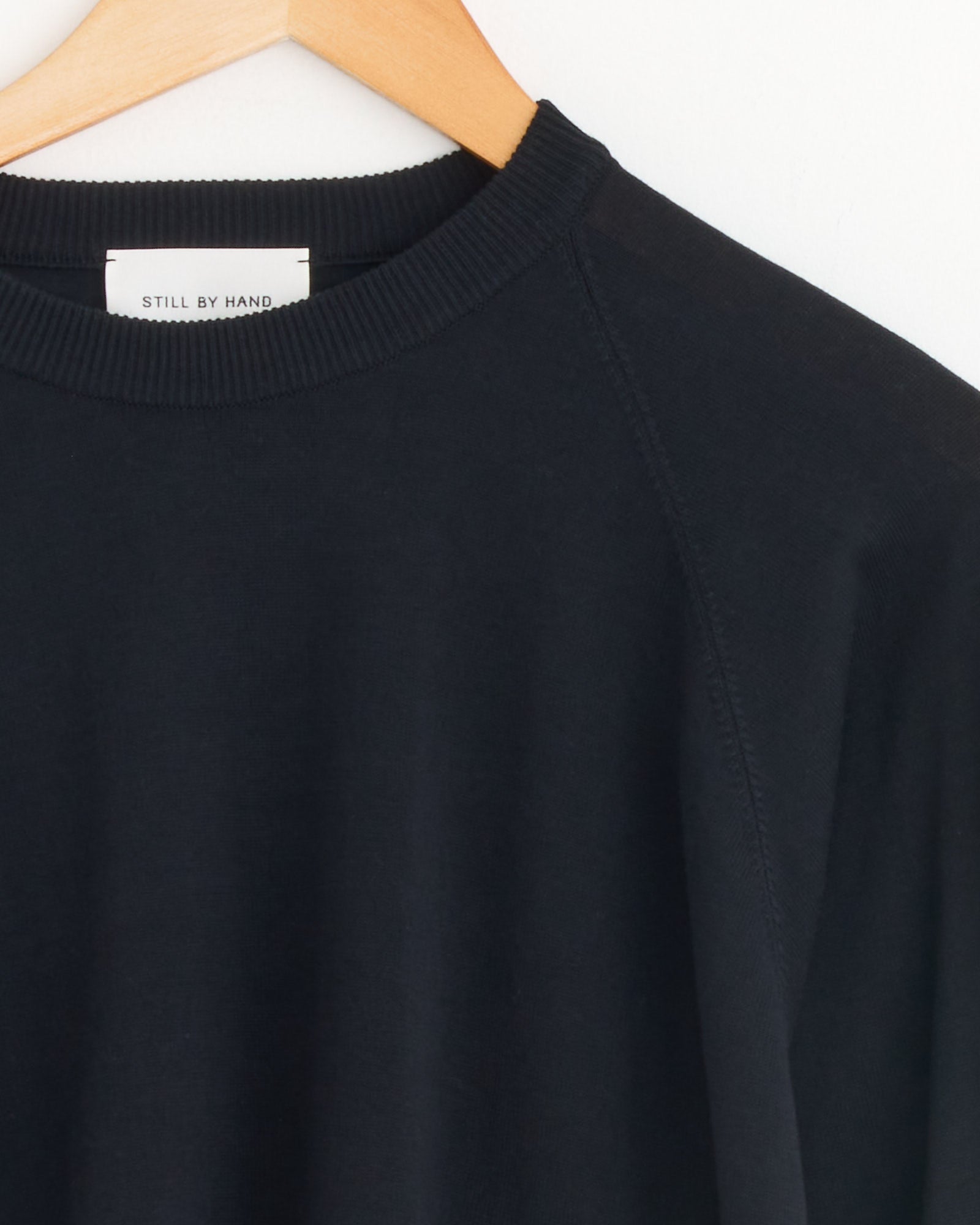 A close-up of the Cotton Silk Pullover in Black Navy by Still By Hand, made in Japan, hanging on a wooden hanger against a white background, shows its ribbed crewneck and a white brand label.