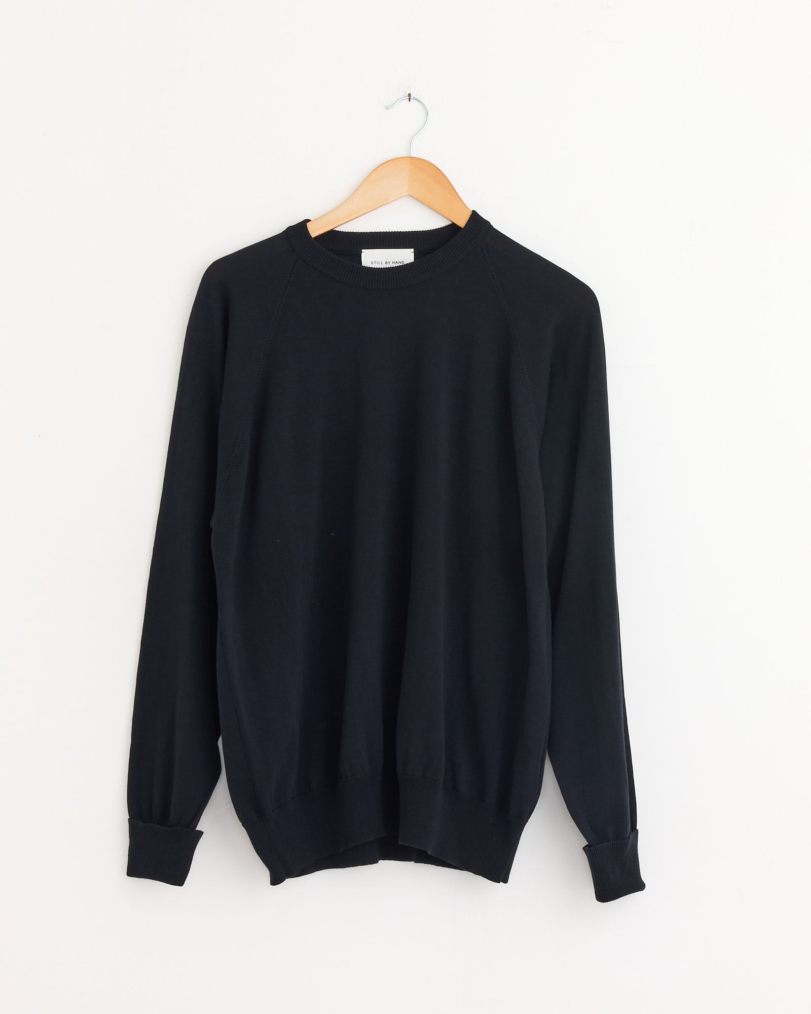 The Cotton Silk Pullover in Black Navy by Still By Hand hangs on a wooden hanger against a white wall, highlighting its lightweight Japanese cotton-silk blend.