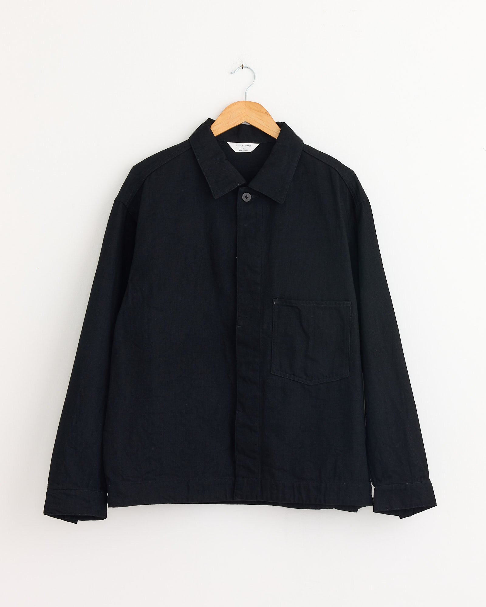 The Still By Hand 12oz Denim Jacket in Black hangs on a wooden hanger against a white wall, combining style and durability with its sturdy 12oz denim construction.
